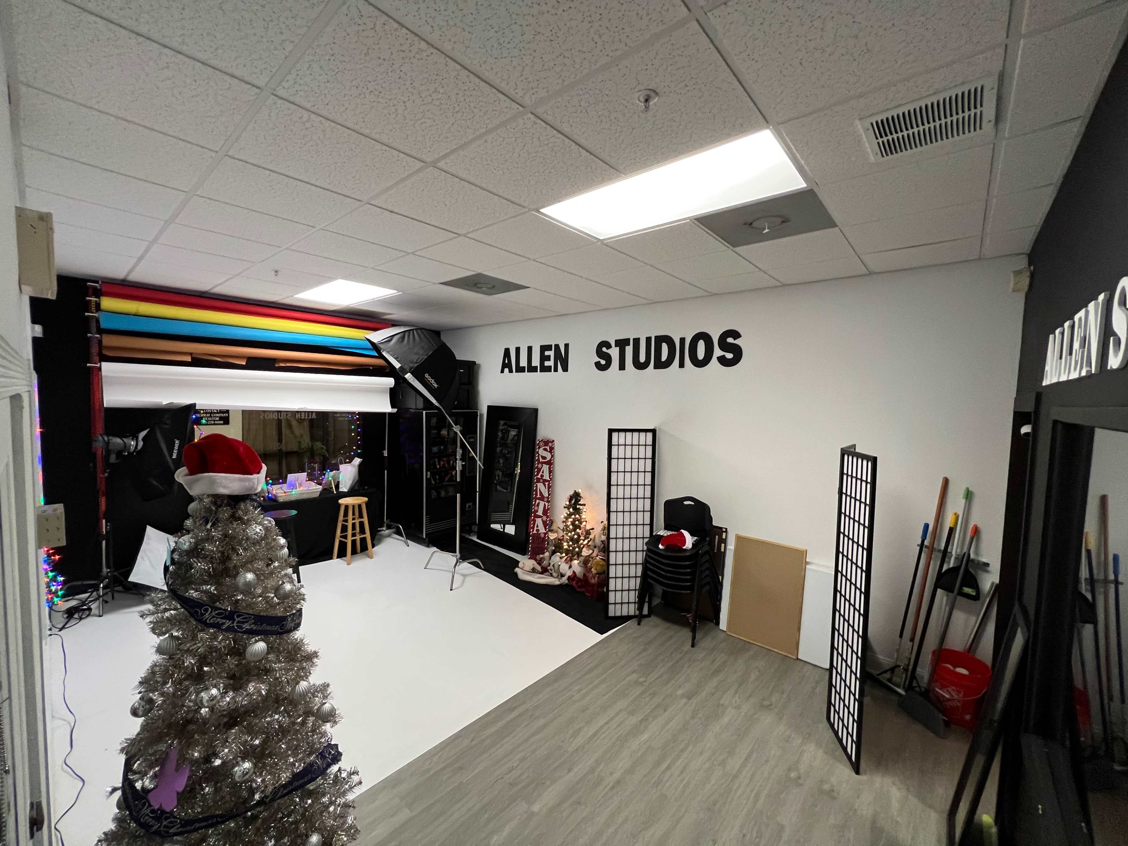 An indoor photography studio featuring a white backdrop, equipment, decorative trees, and the words "ALLEN STUDIOS" on the wall.
