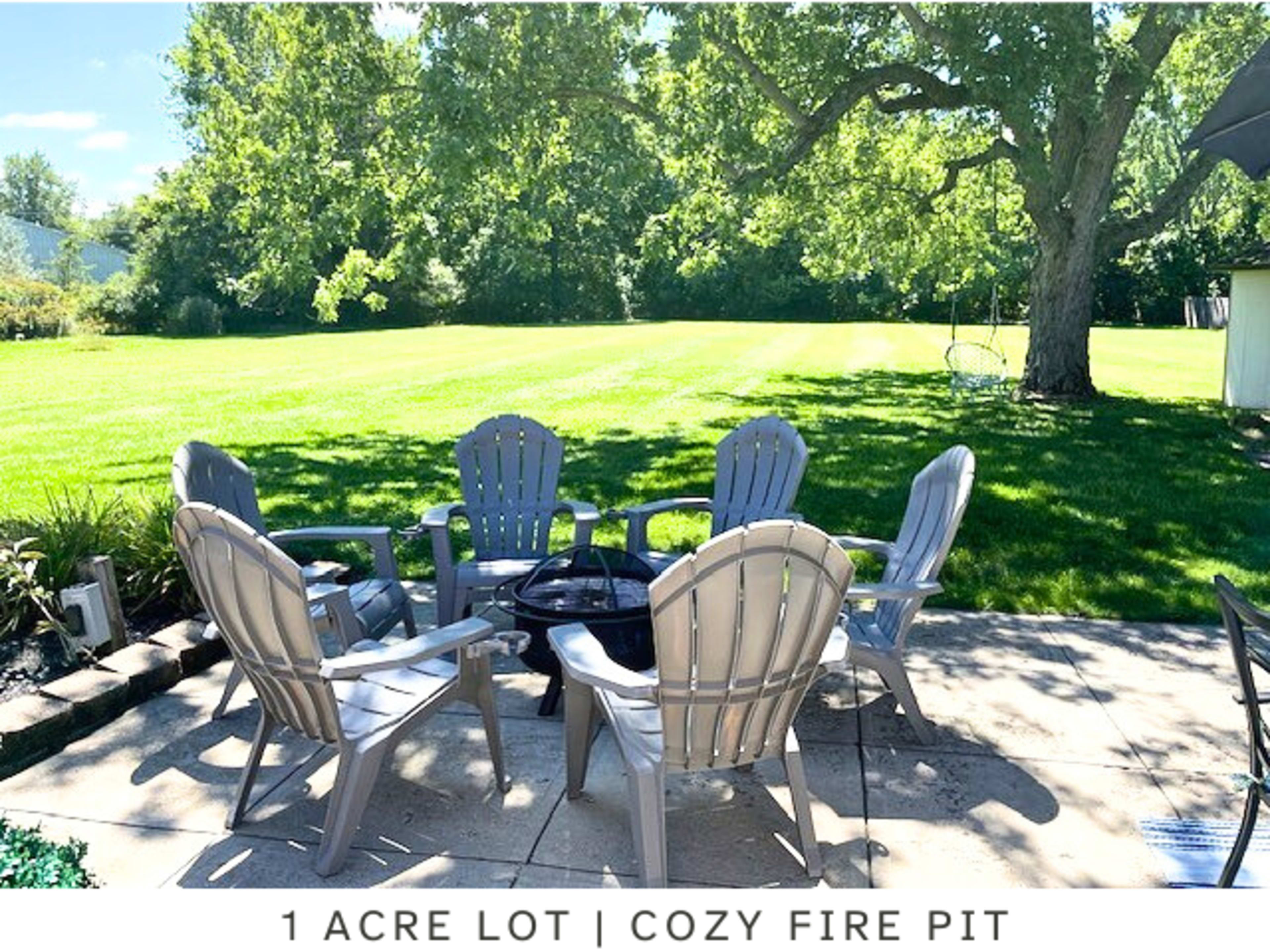 A spacious backyard with a circular arrangement of six gray chairs around a fire pit on a stone patio, set in a grassy one-acre lot.