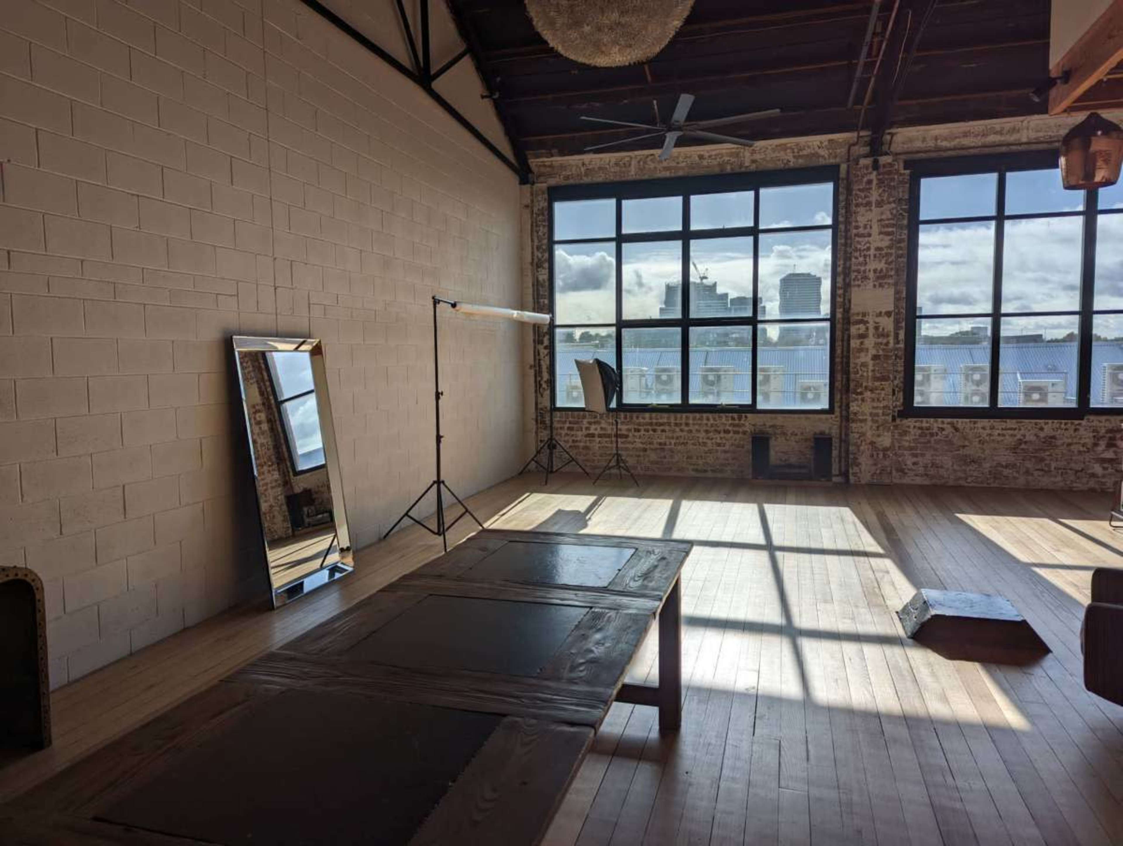 The image shows a spacious, well-lit room with large windows, a wooden table, a mirror, and a lighting stand in the corner.