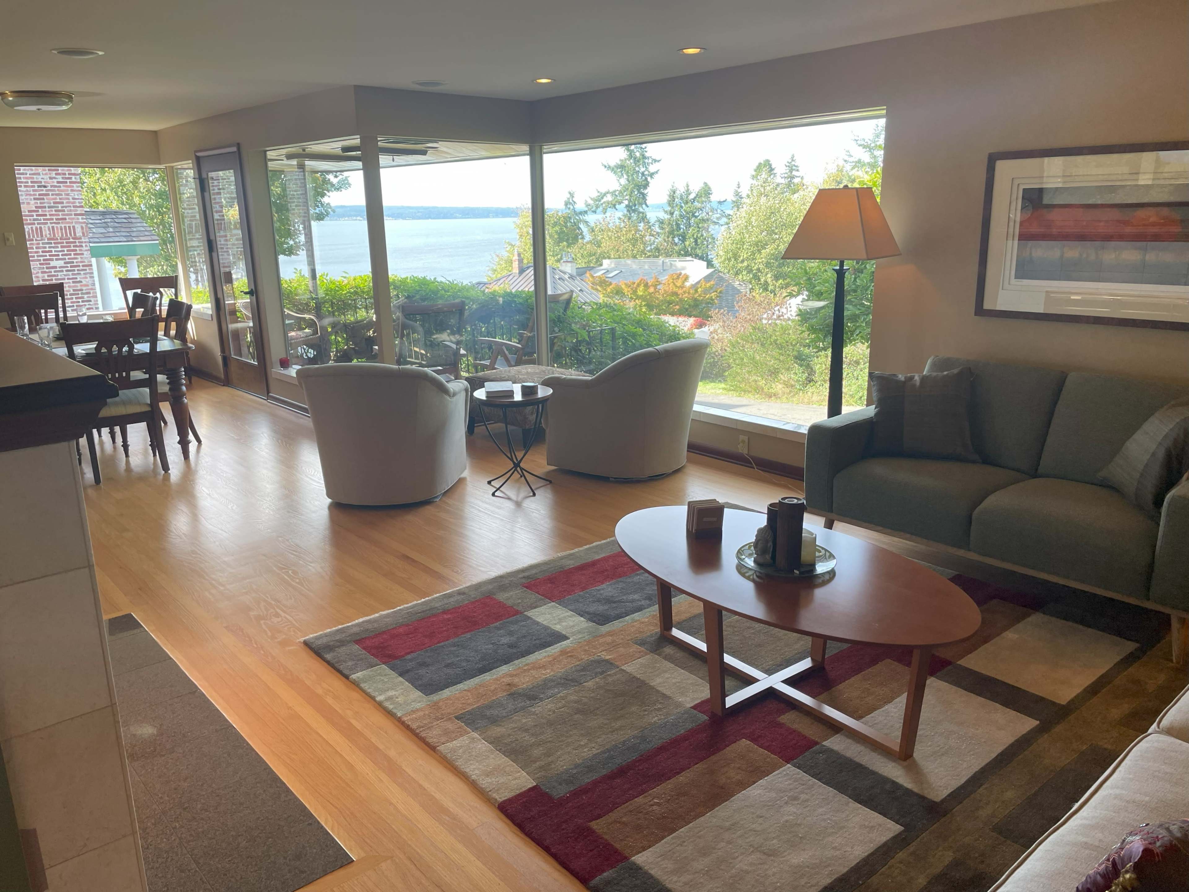 The living room features a couch, chairs, and a coffee table, with large windows overlooking a scenic view of trees and water.