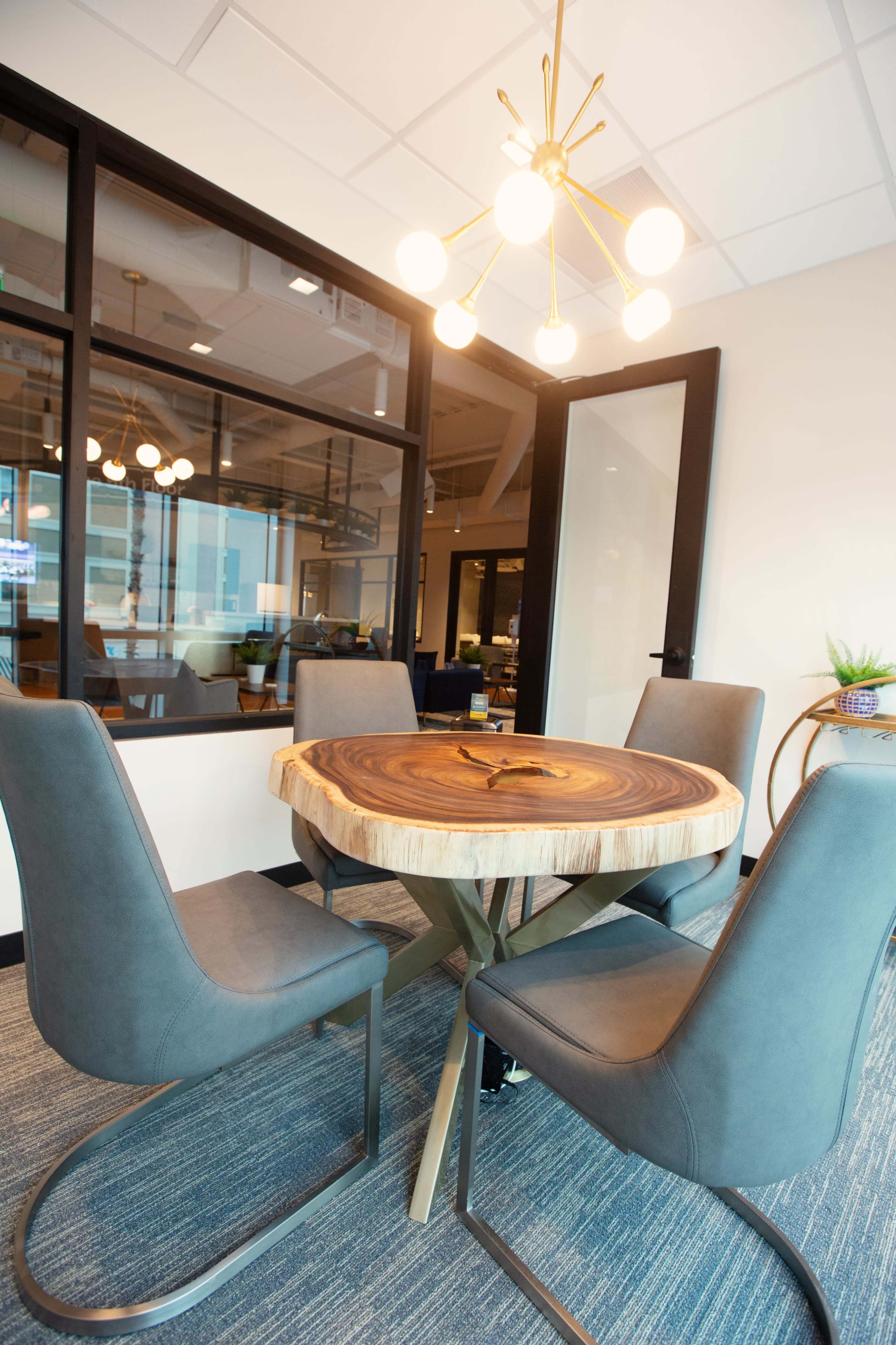 A round wooden table with four upholstered chairs sits in a modern conference room, illuminated by a contemporary light fixture.