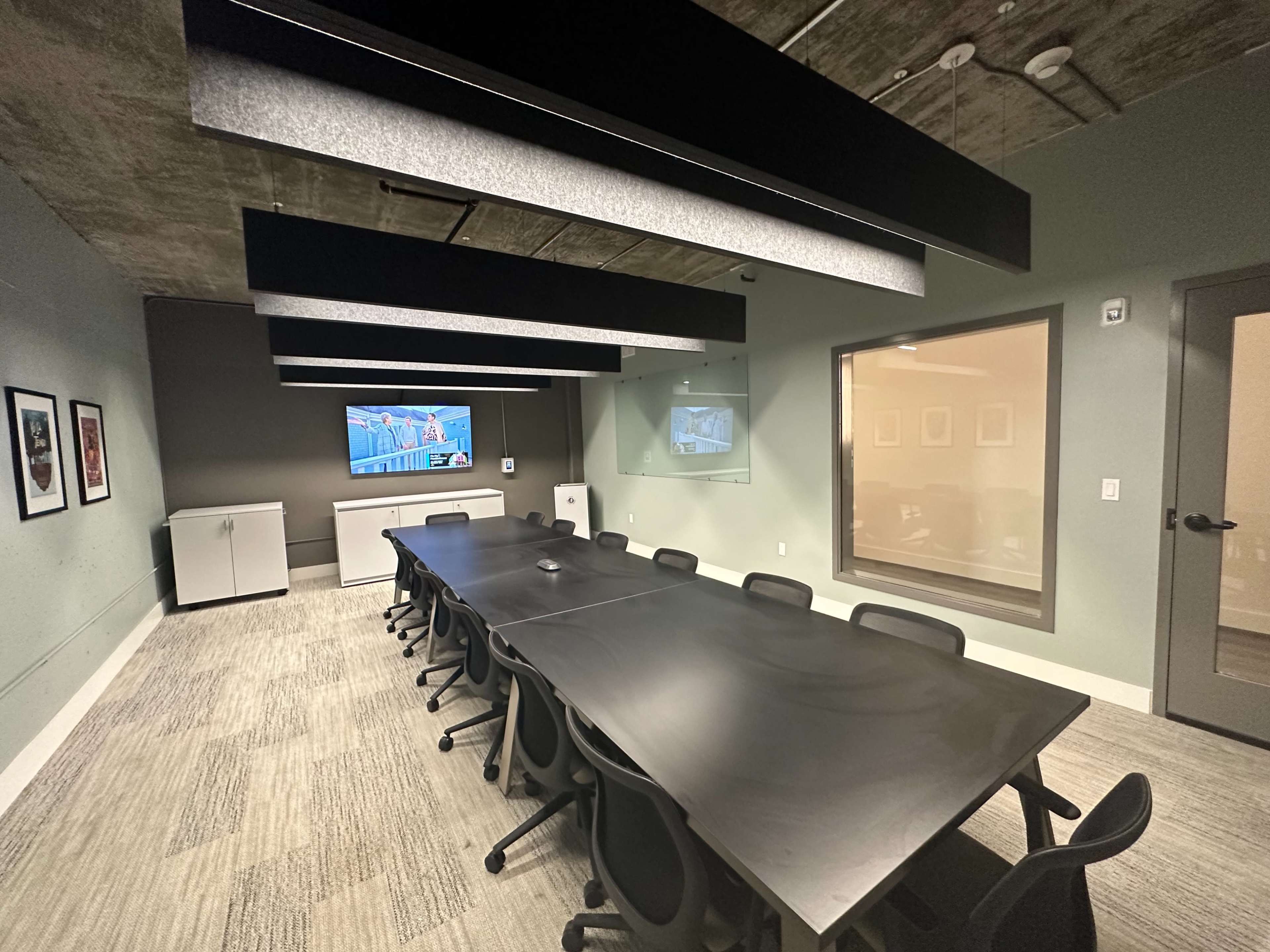 A modern conference room features a long black table surrounded by chairs, with a large screen and a glass-enclosed area for video presentations.