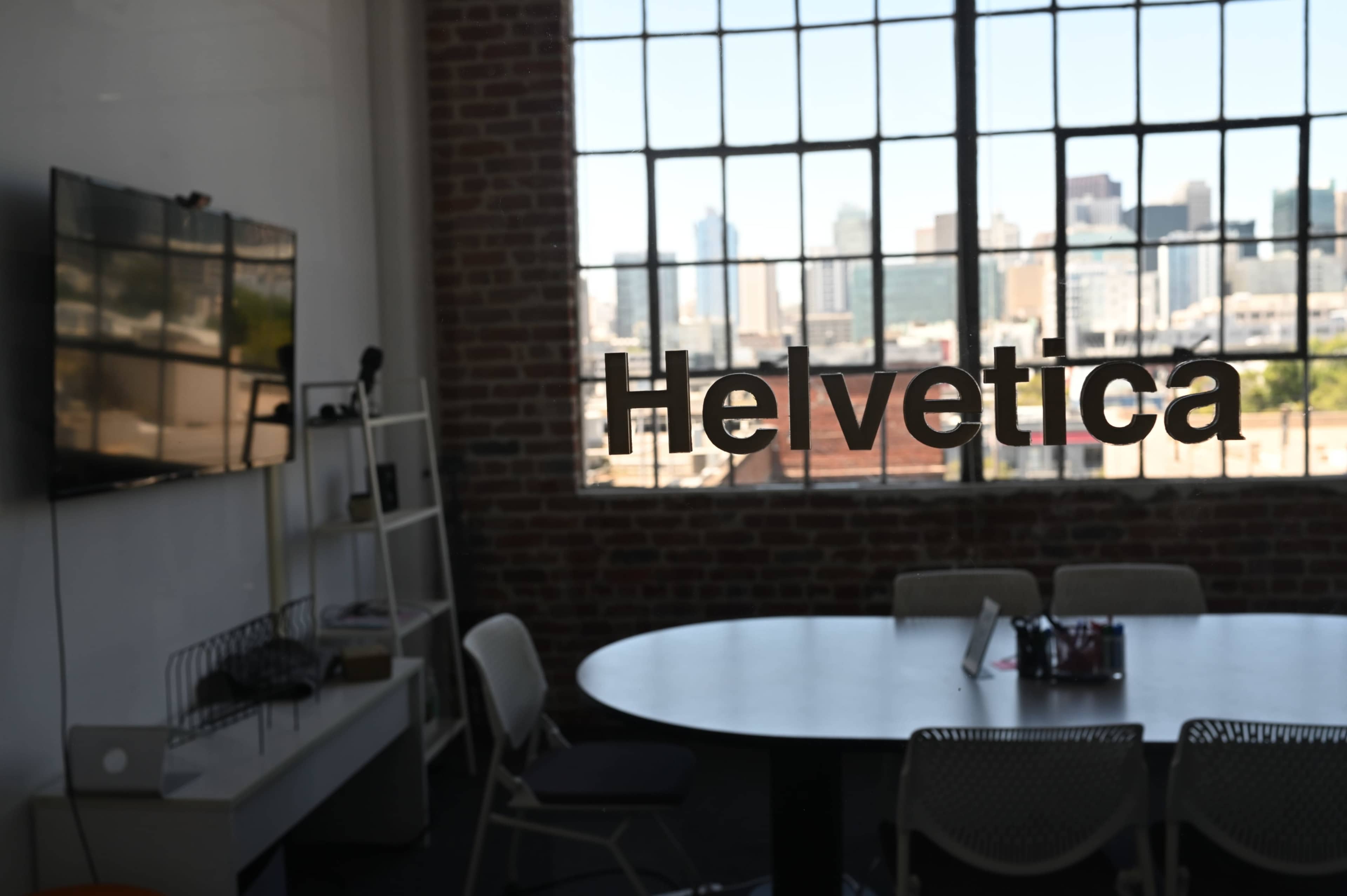 The image shows a modern conference room with a round table, a television on the wall, and large windows displaying a city skyline in the background.