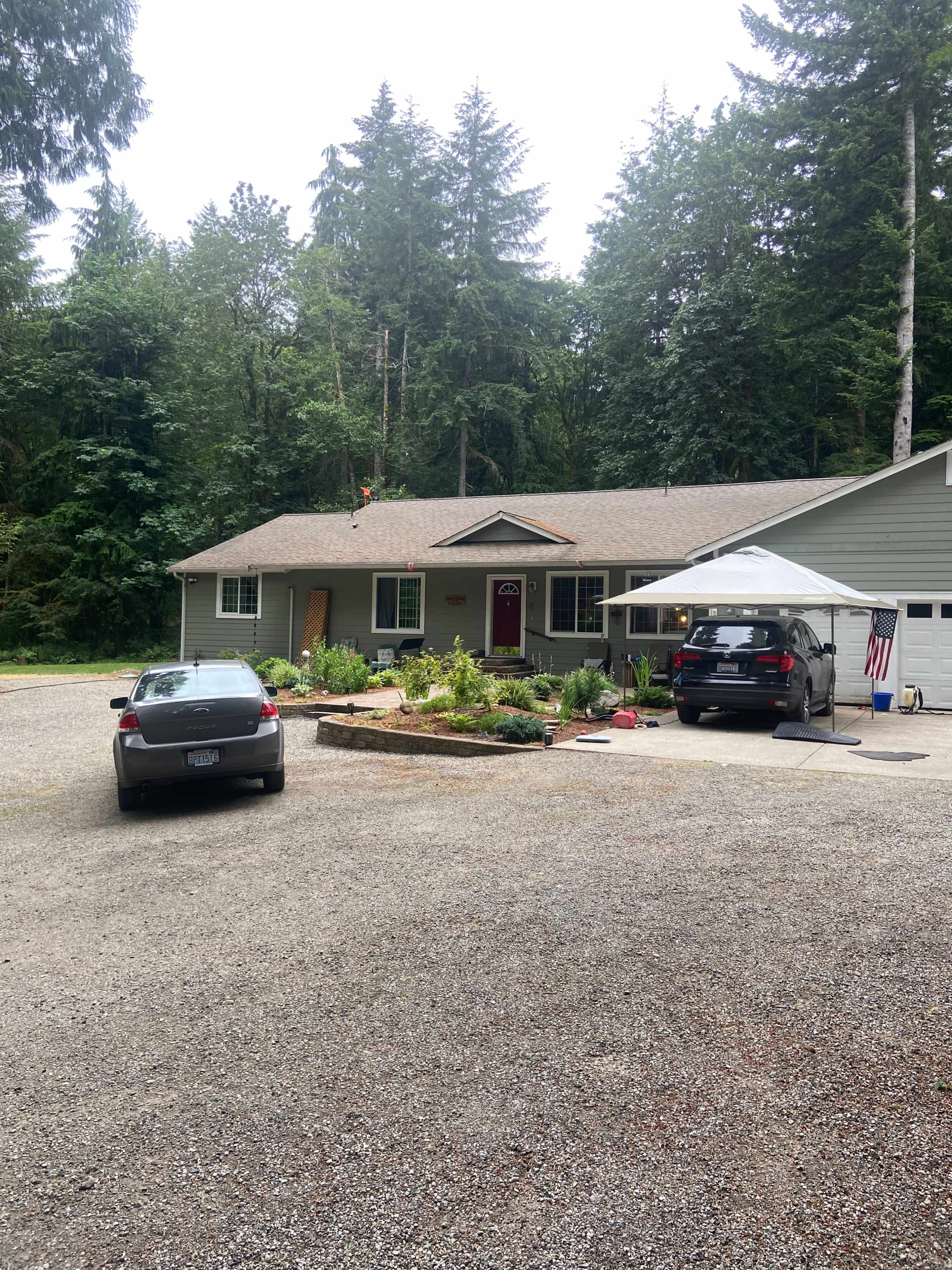 A single-story, light-colored house with a gravel driveway, surrounded by greenery and two parked cars.