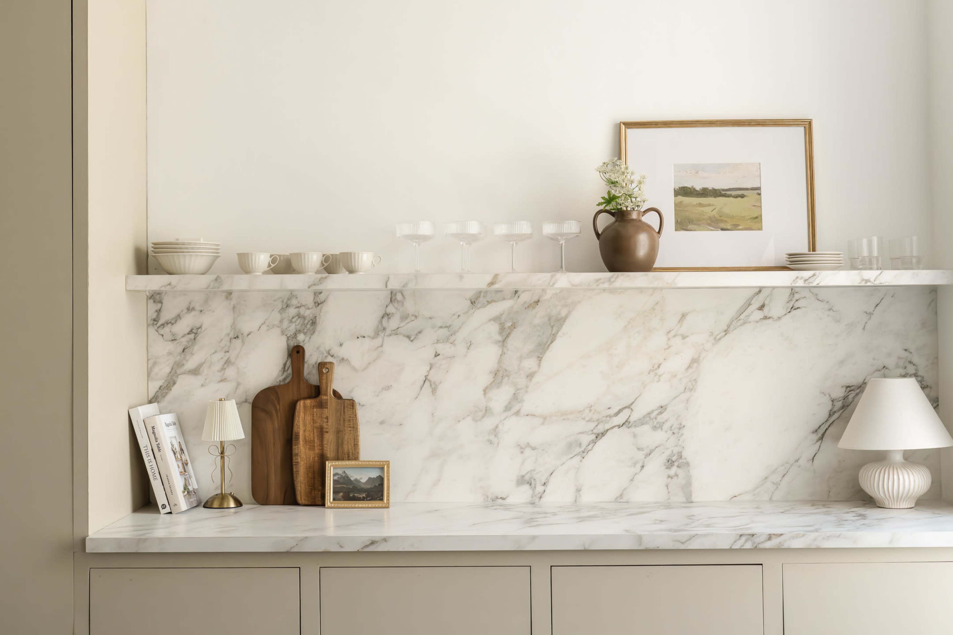 A minimalist shelf displays dinnerware, glasses, and decorative items against a marble backdrop.