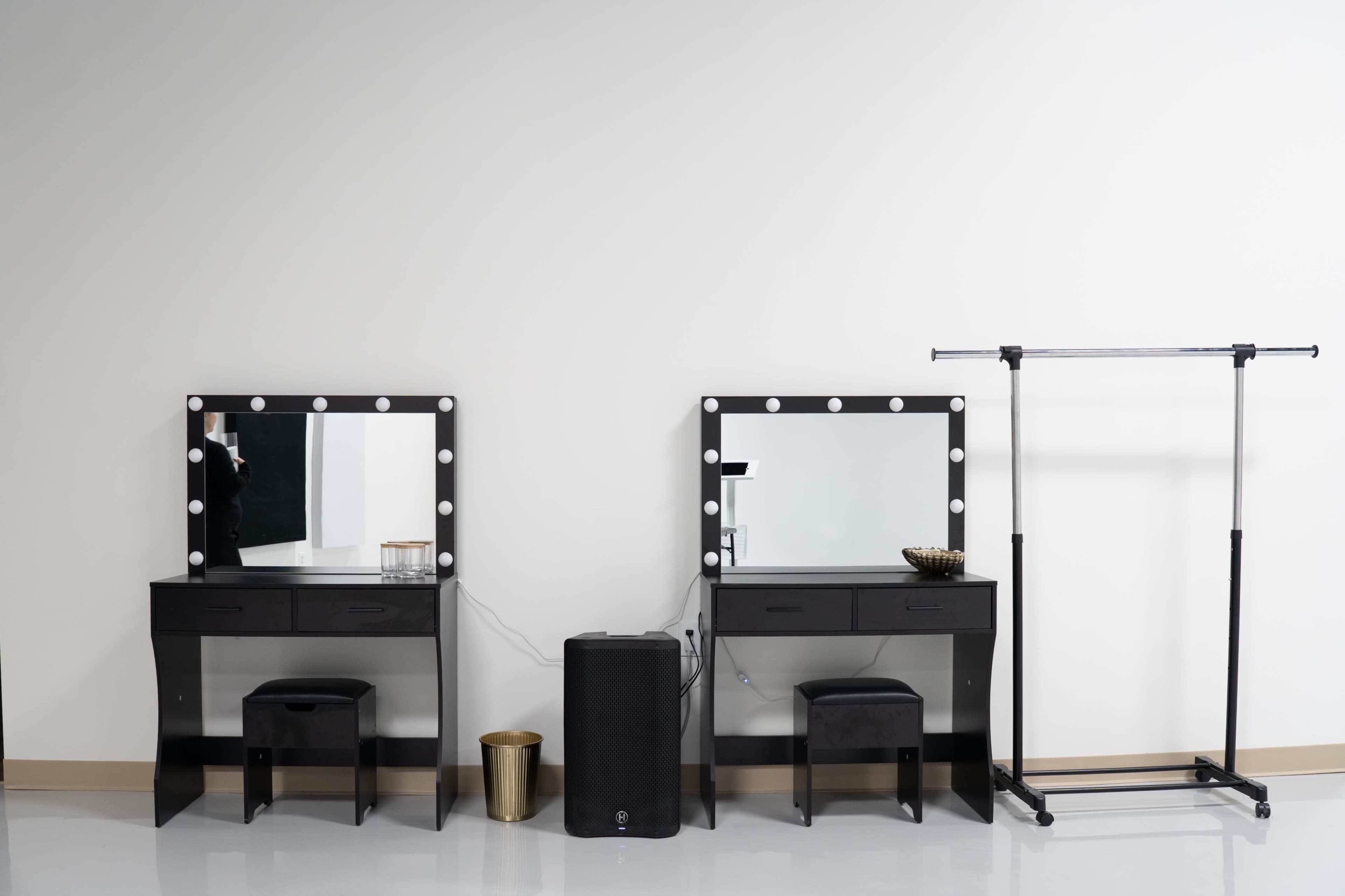 The image features two identical black vanity tables with mirrors and lights, positioned beside a clothing rack and a small trash can on a smooth floor.