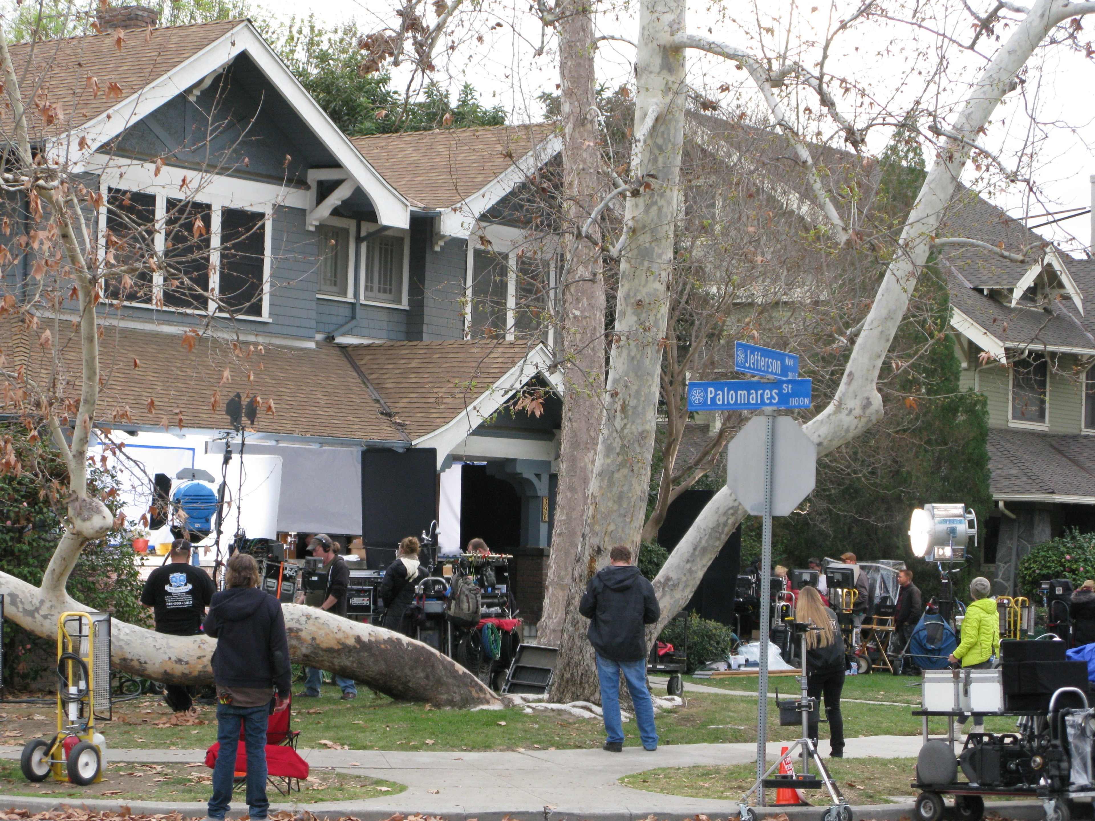 A film crew sets up equipment outside a house with a sign for Palomares Street in view.