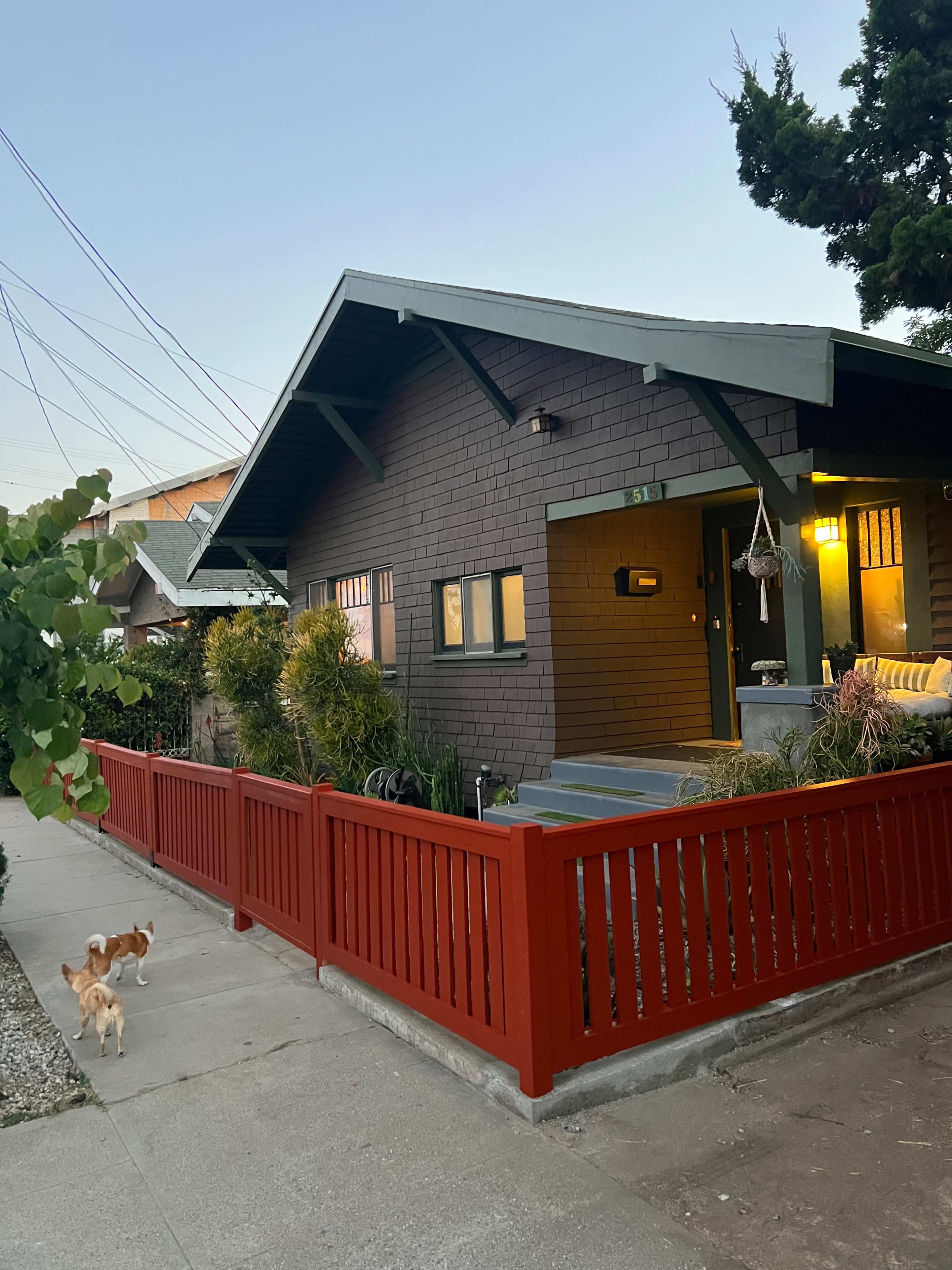 A single-story house with a dark exterior and green trim is located on a sidewalk bordered by a red fence, with two small dogs walking nearby.