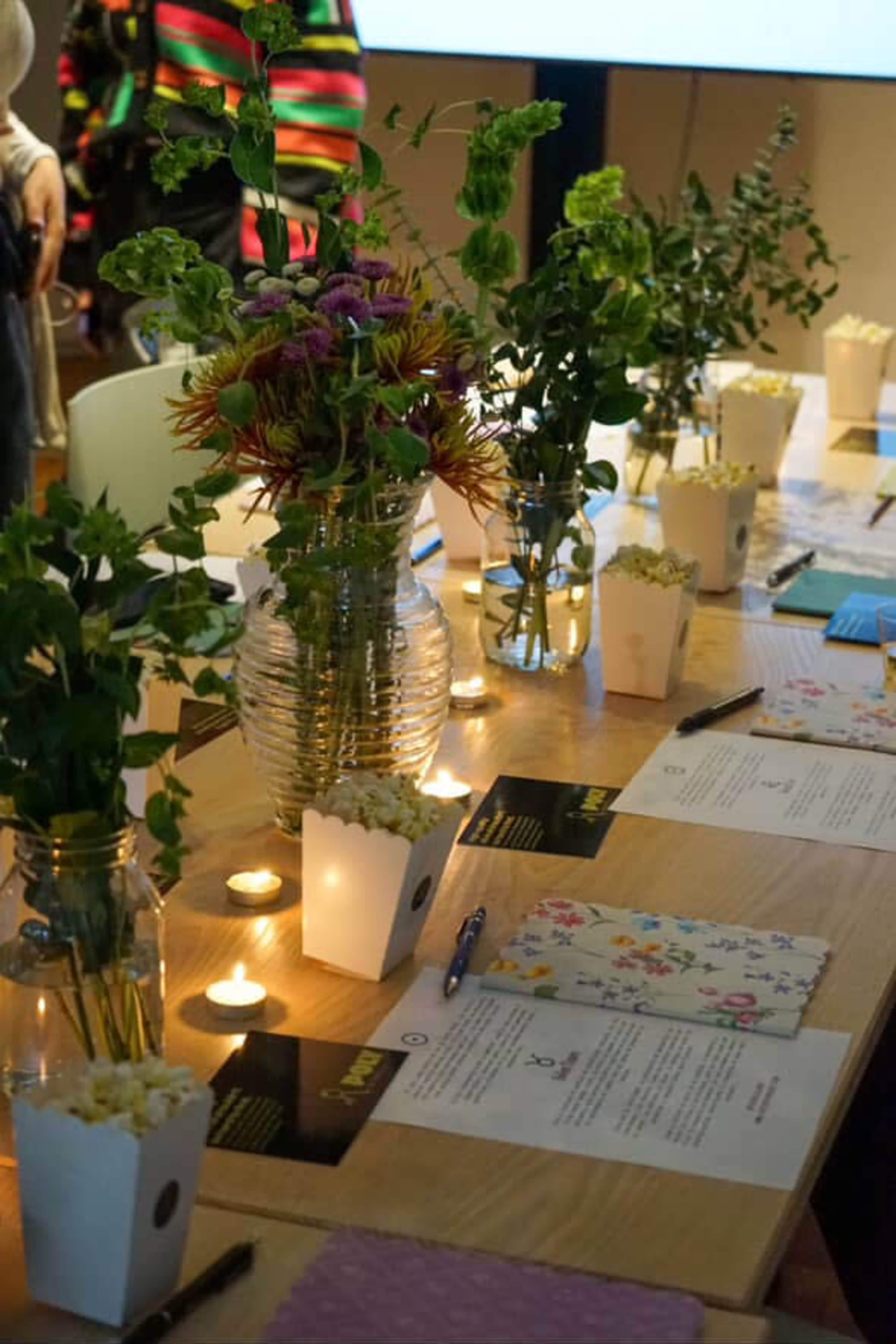 A long wooden table is set with floral arrangements in vases, small containers of popcorn, candles, and neatly arranged notepads and pens.
