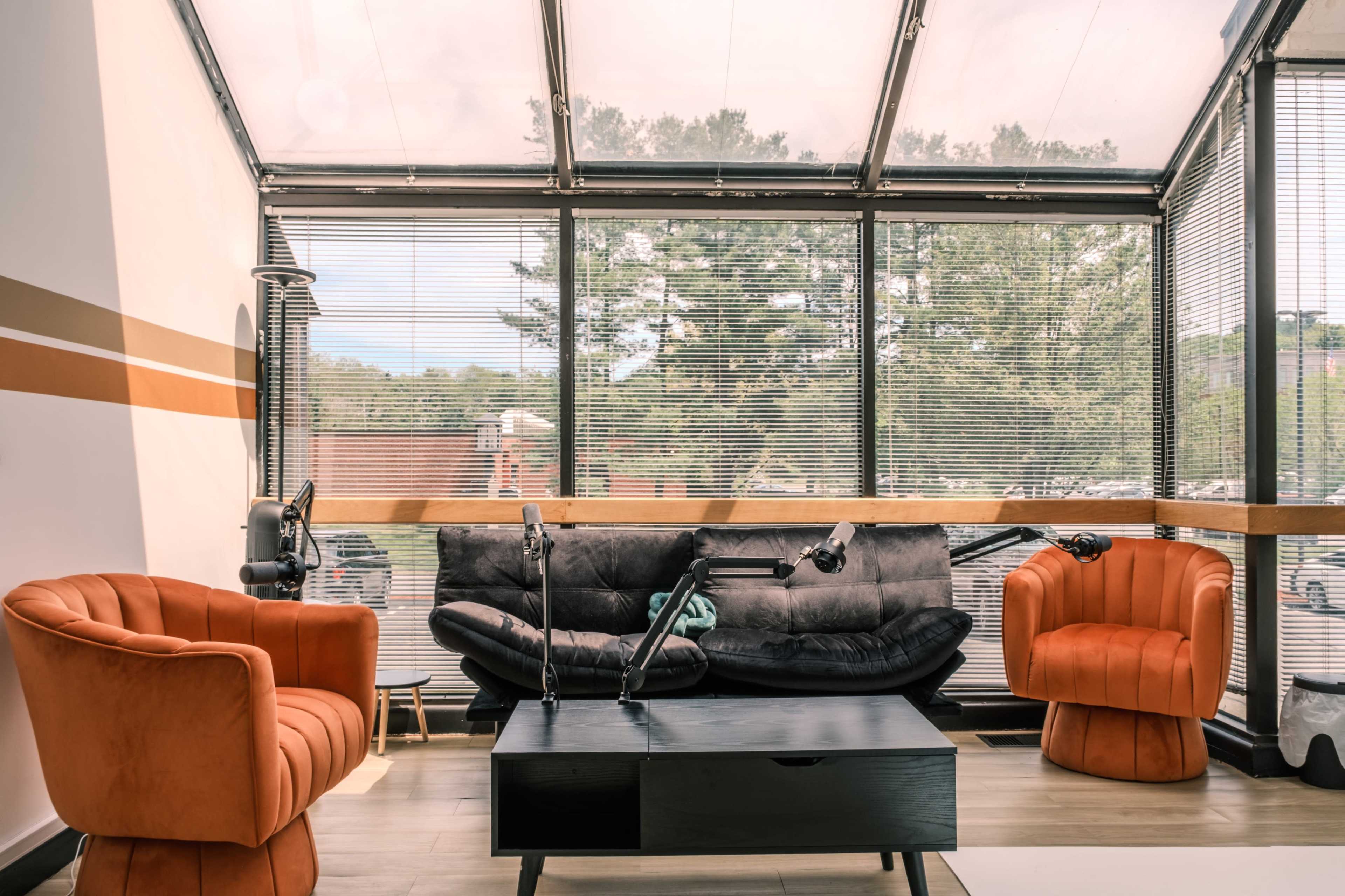 The image shows a modern seating area with an orange armchair on each side of a black coffee table and a gray couch in a sunlit room featuring large windows with blinds.