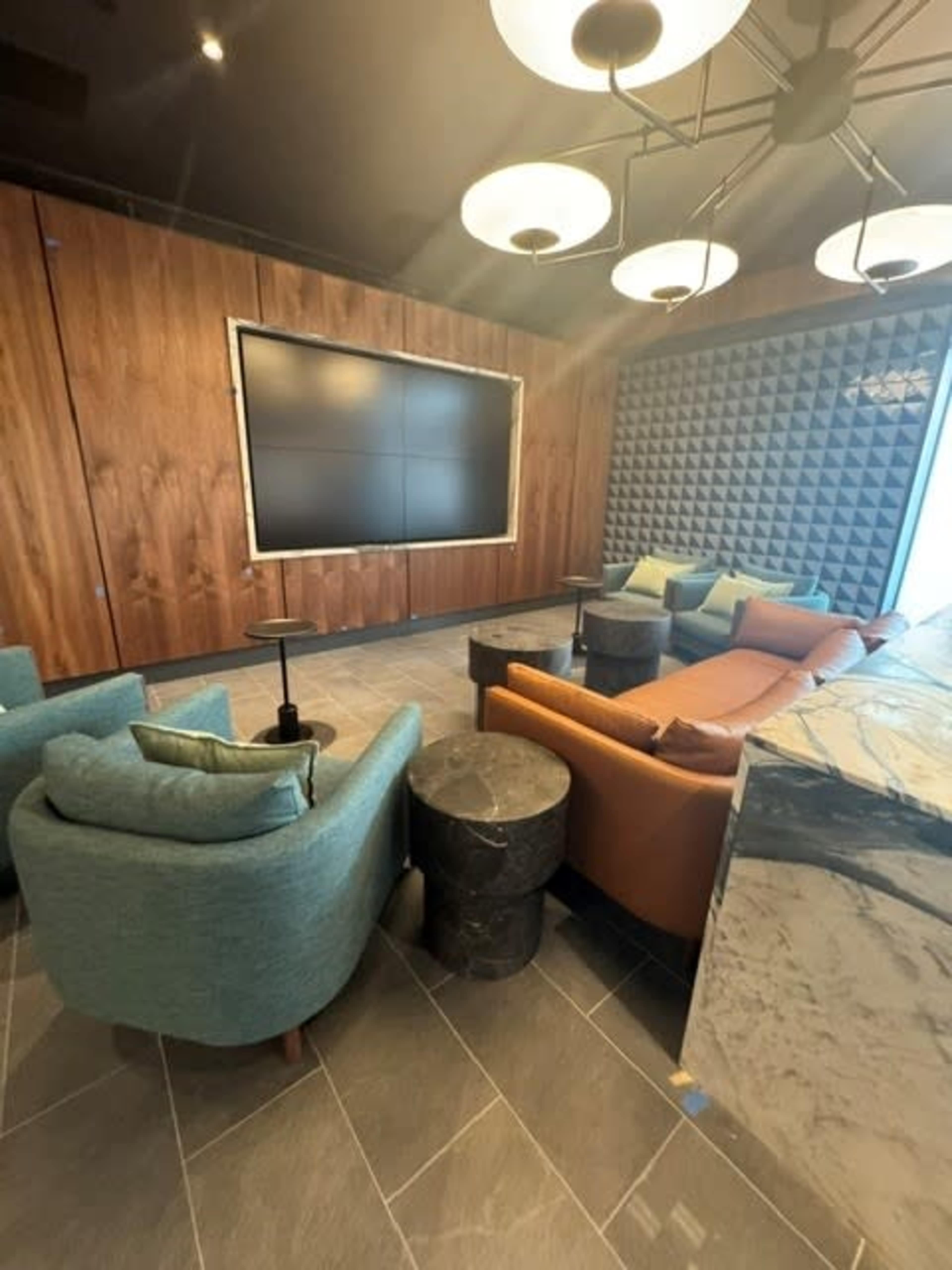 The image shows a modern lounge area with two distinct seating arrangements, featuring a large screen on one wall and textured panels on another.