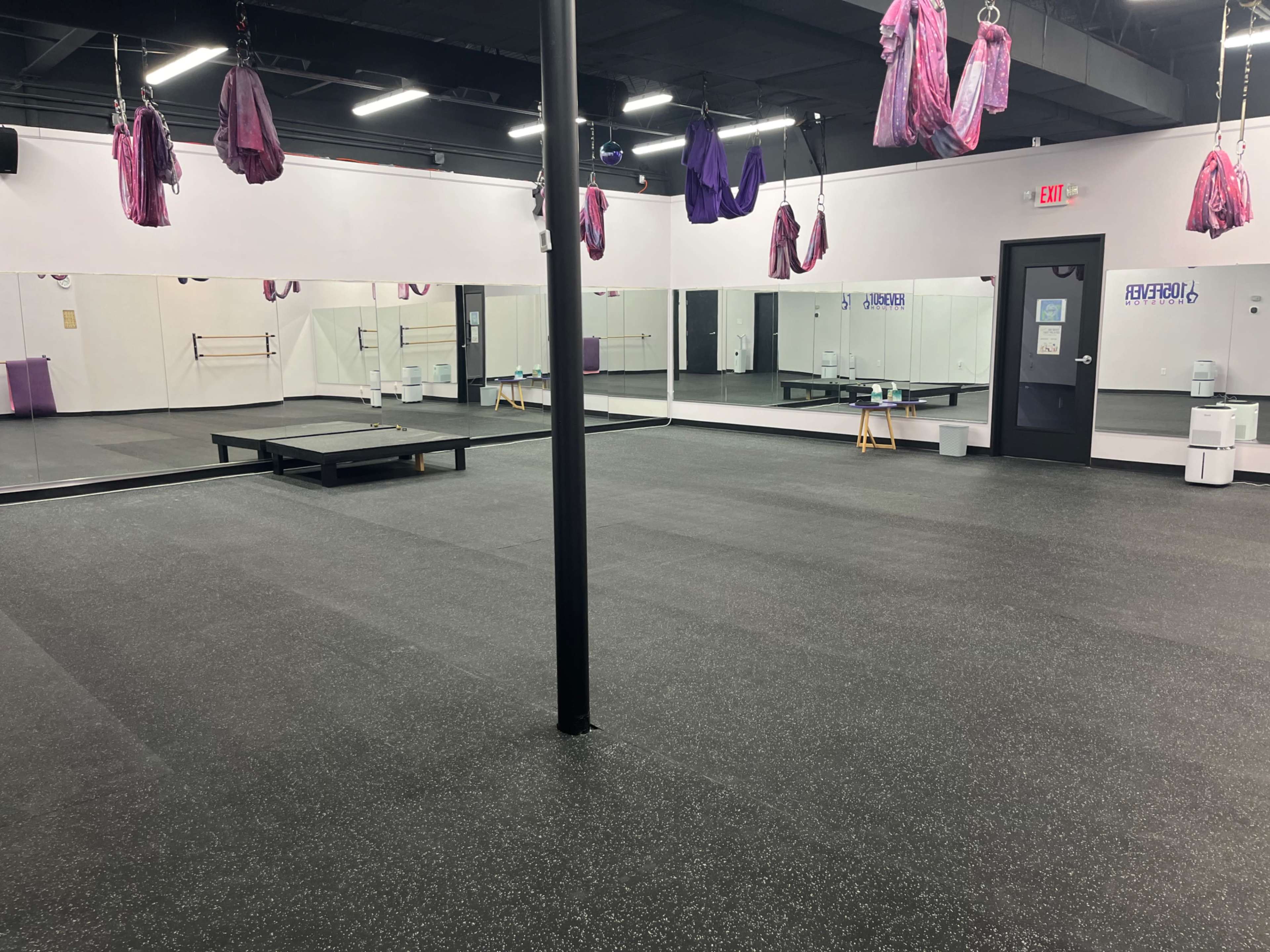 An empty fitness studio with a black rubber floor, a mirrored wall, and hanging exercise equipment from the ceiling.