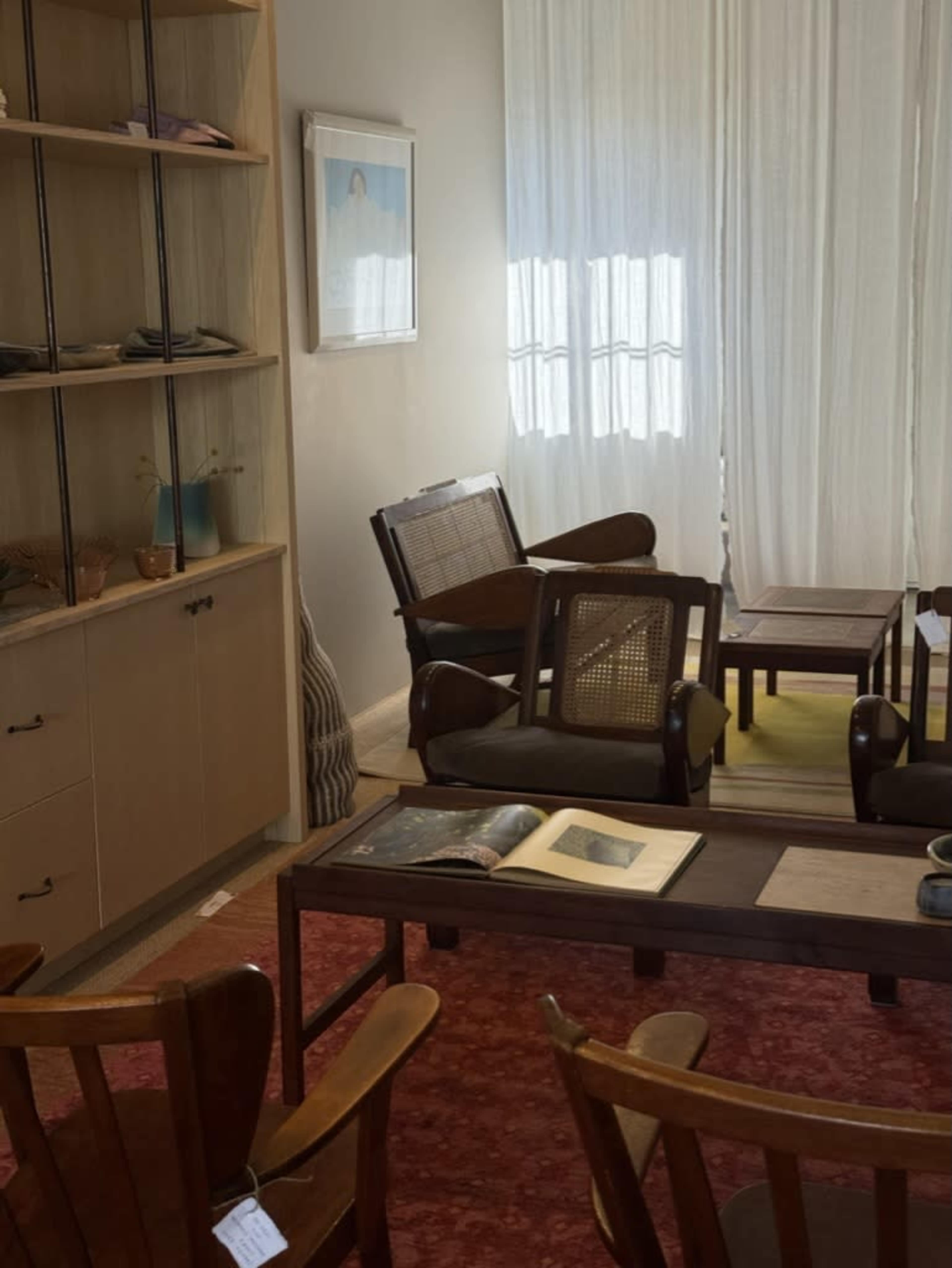 The image shows a cozy interior with wooden furniture, shelving containing decorative items, and natural light filtering through sheer curtains.