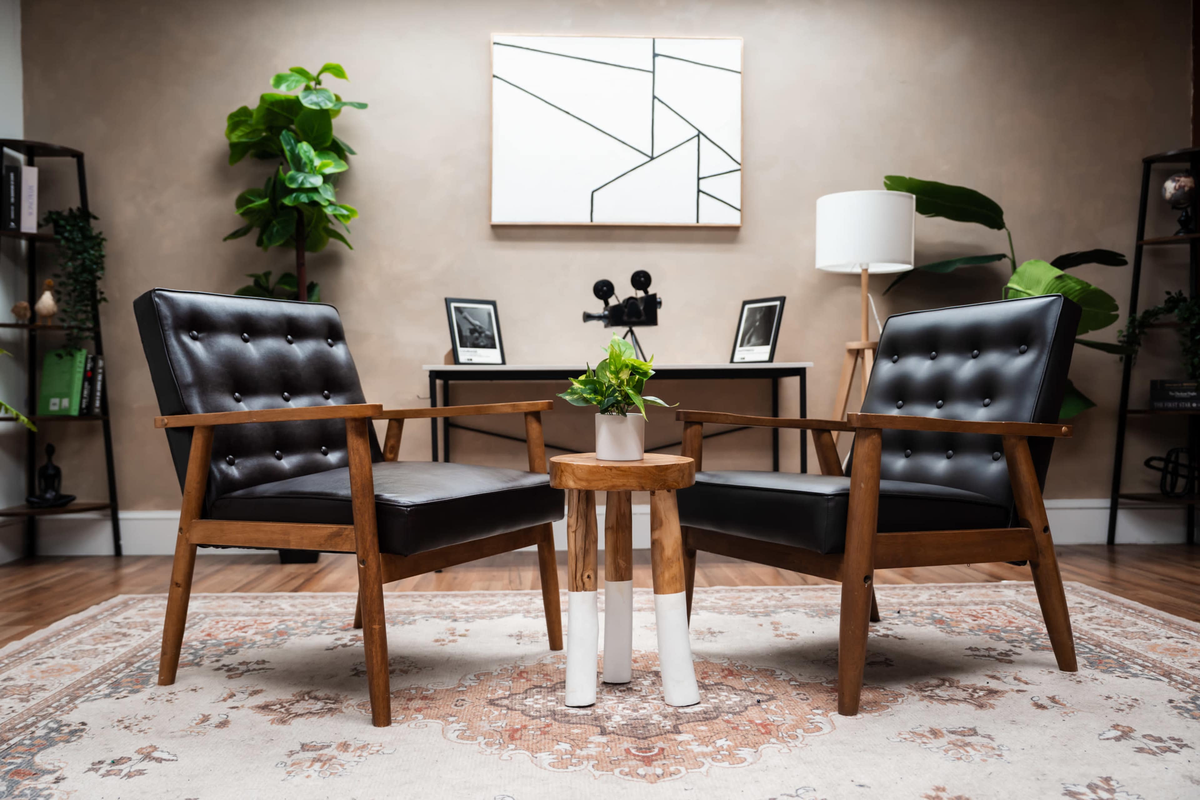 The image shows a minimalist interior design featuring two black mid-century armchairs facing each other, a small wooden side table with a potted plant, and a desk with decorative items in the background.