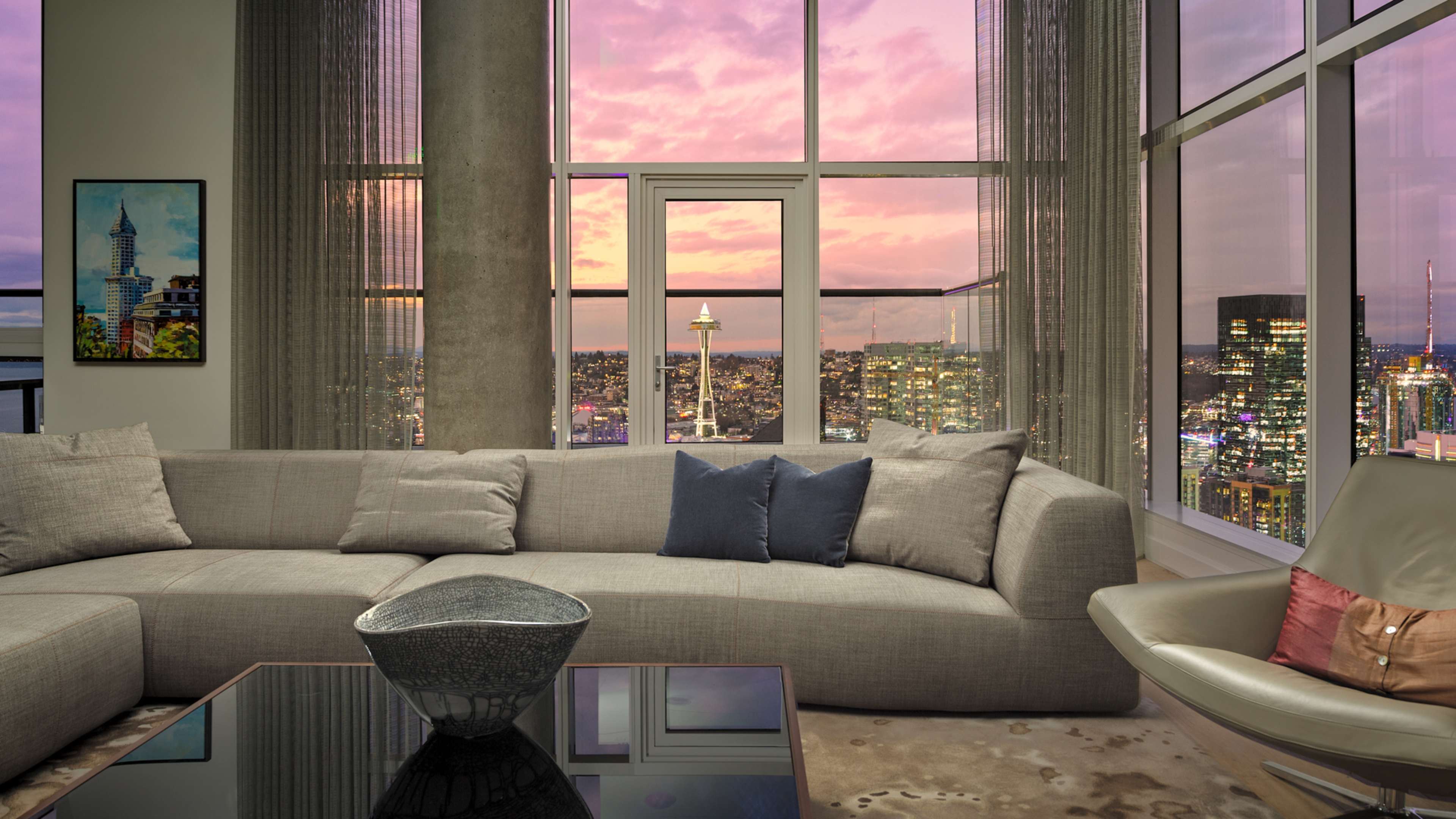 A modern living room features a large sectional sofa, a coffee table, and expansive windows showcasing a city skyline at sunset.