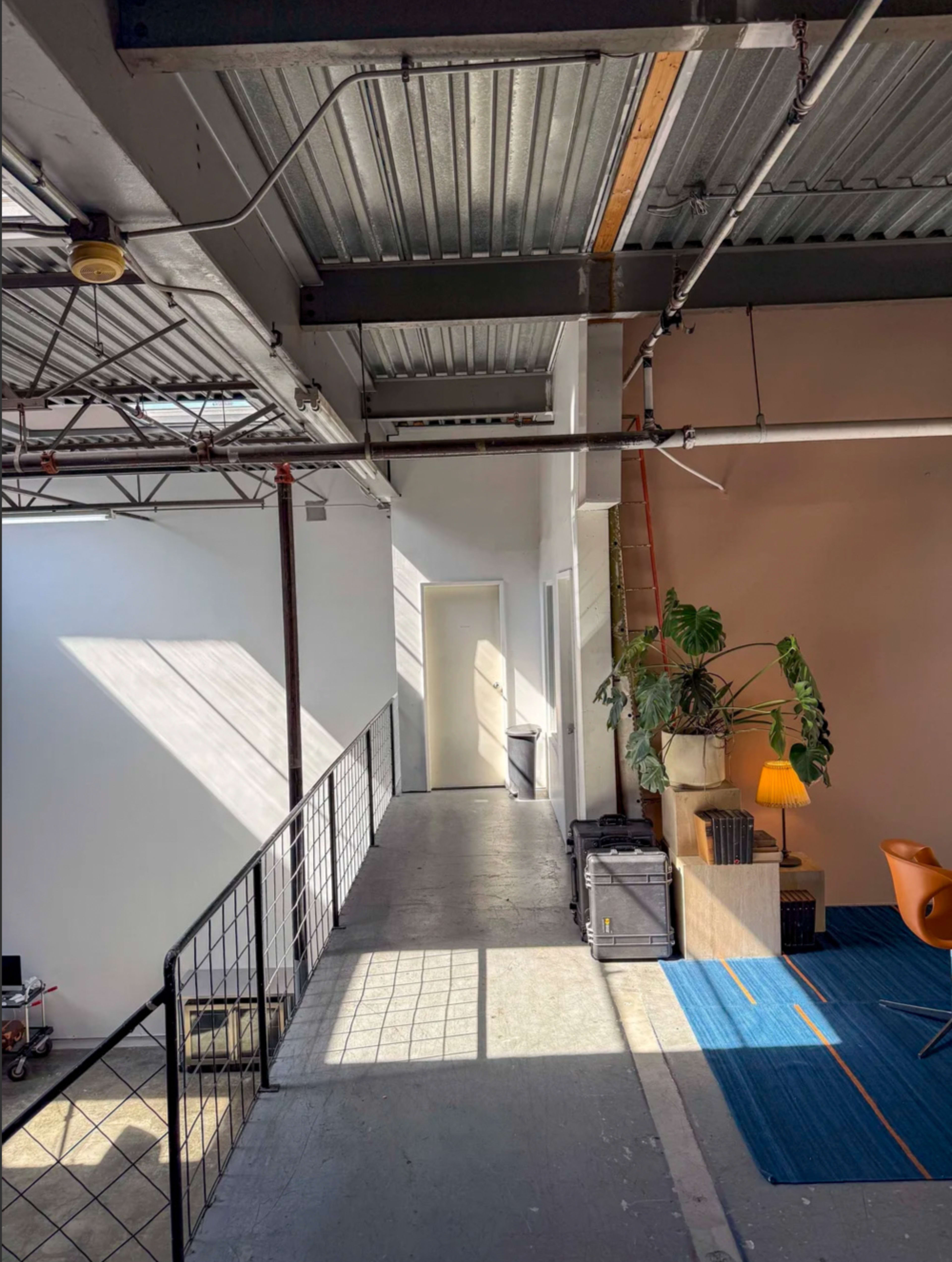 The image shows a modern office space with a long hallway featuring bright sunlight streaming through a window, leading to a door and a small seating area with a plant and lamp.