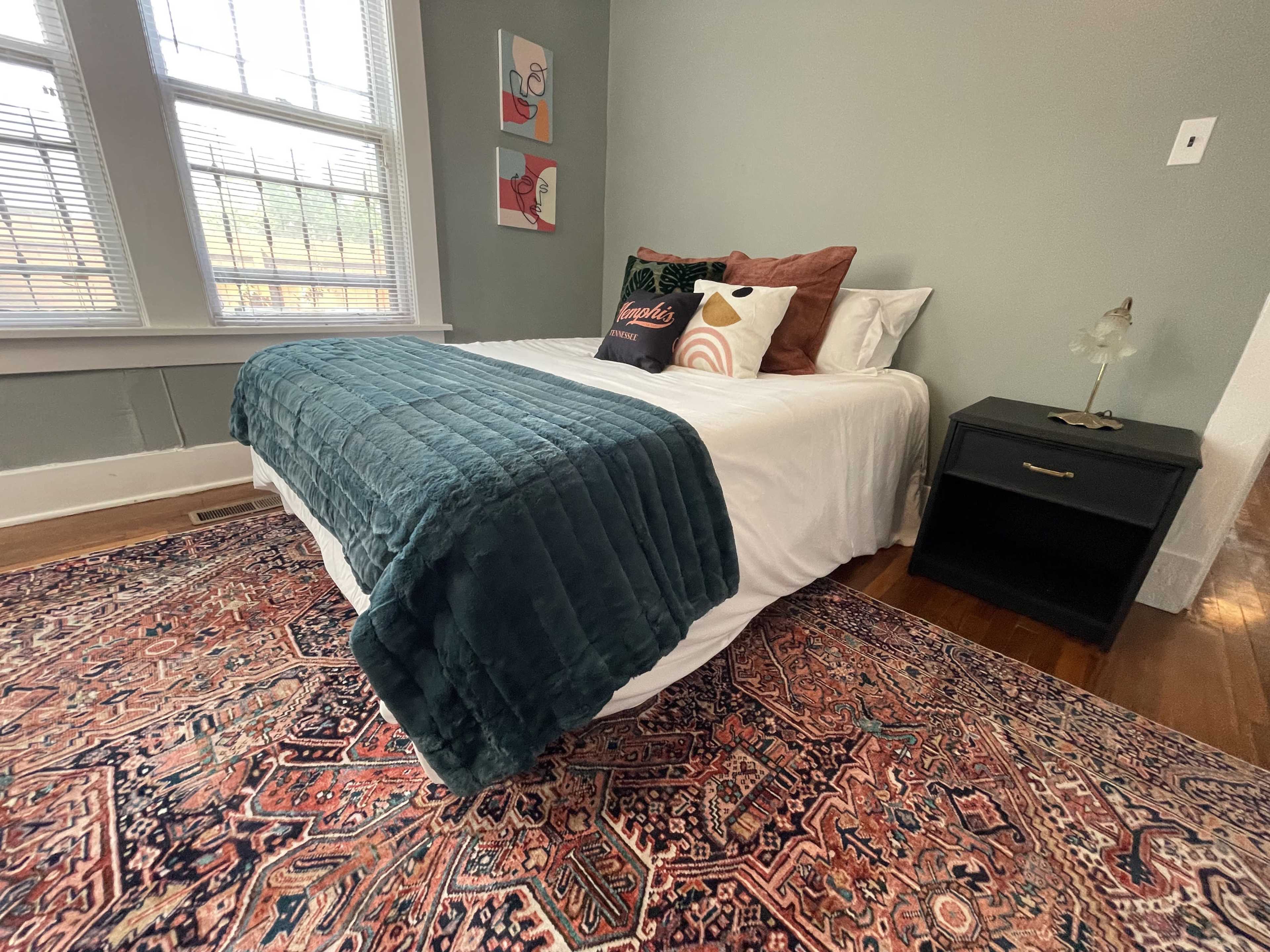 A neatly made bed with a green throw blanket and several decorative pillows, positioned beside a black nightstand in a well-lit room with a large window.