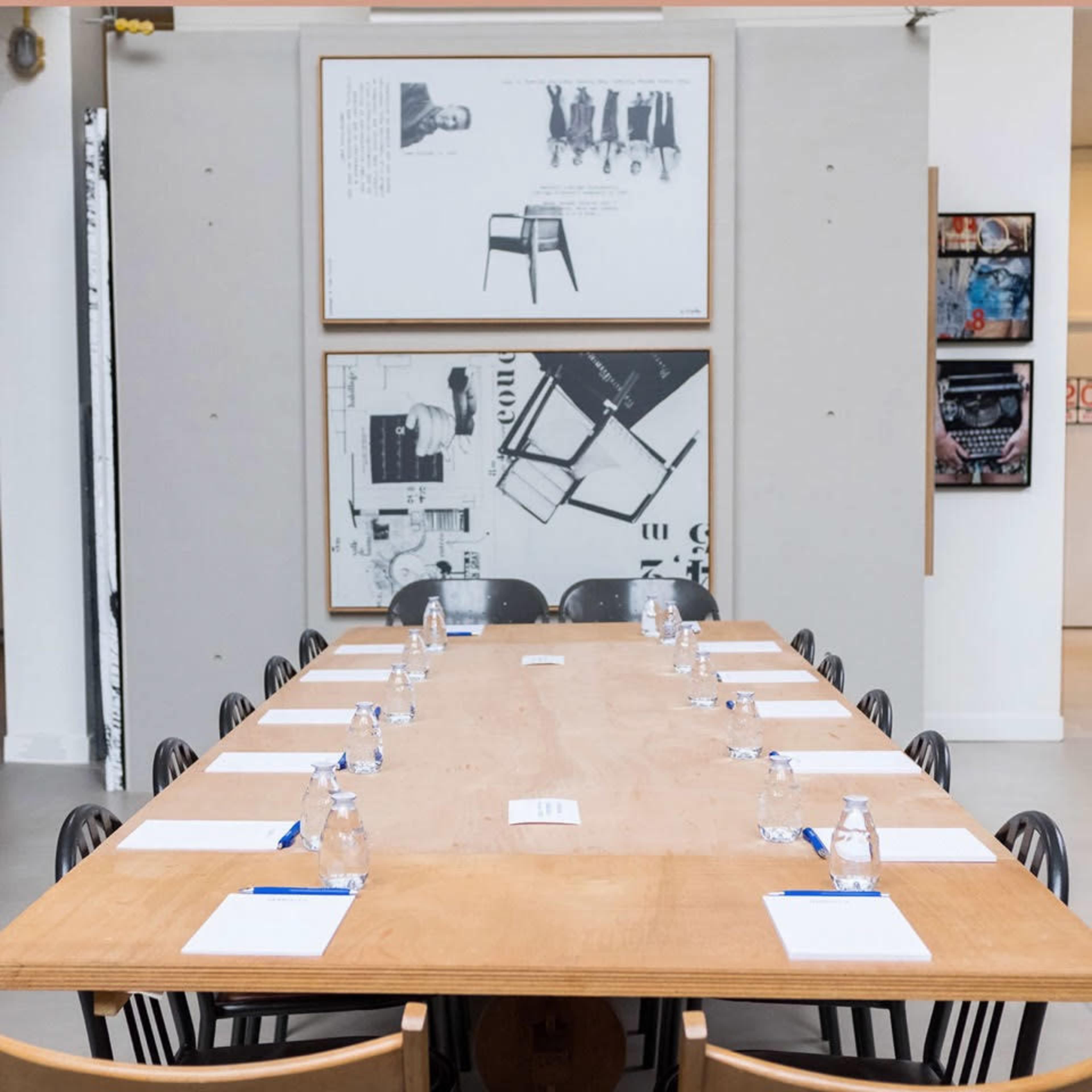 A long wooden conference table is set with water bottles and notepads, surrounded by black chairs in a room featuring framed artwork on the walls.