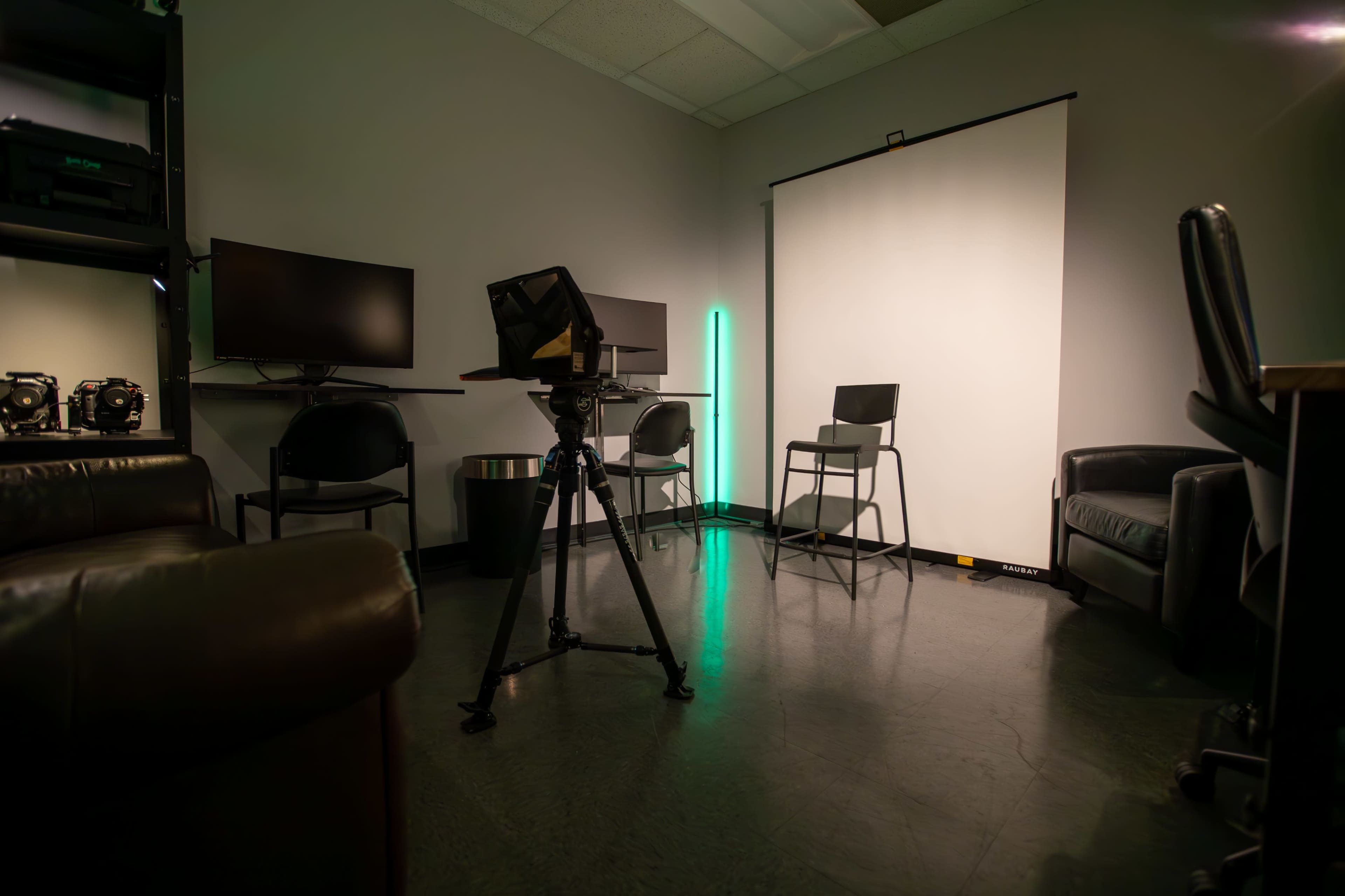 The image shows a small studio space with two monitors, a tripod, a chair, and a white backdrop with a green LED light.
