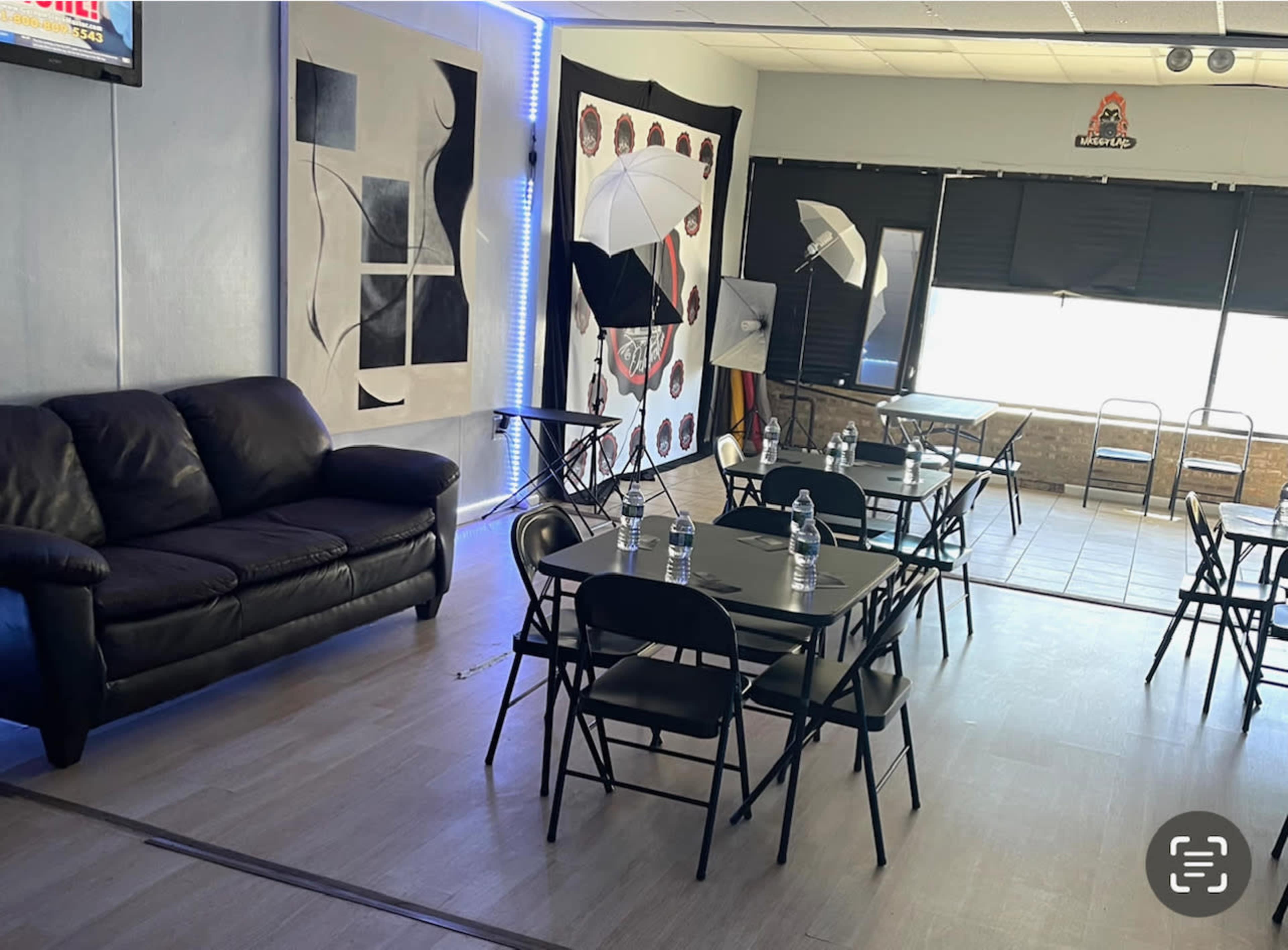 A room with a black couch, multiple folding tables with water bottles, and lighting equipment positioned near the windows.