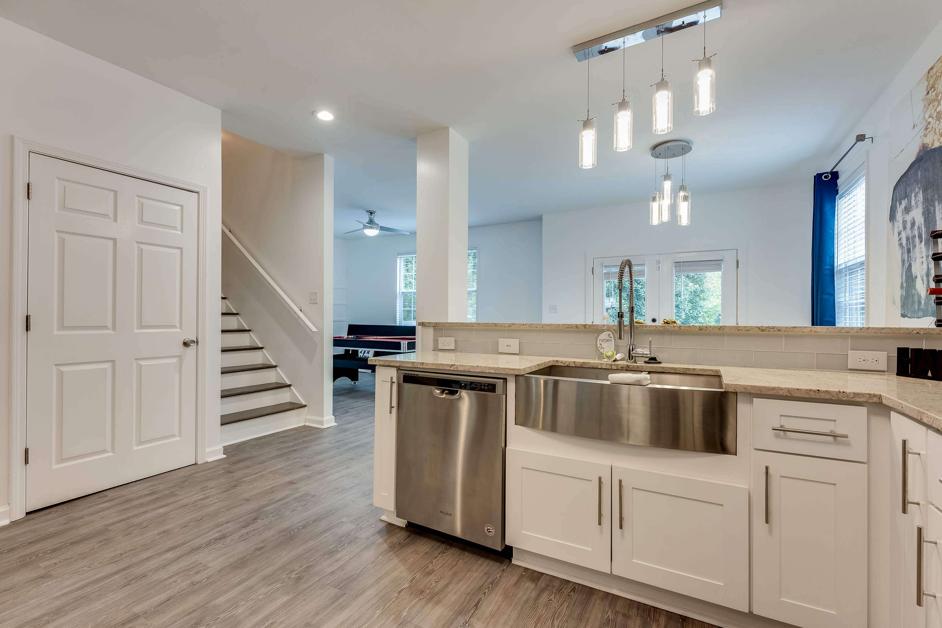 A modern kitchen featuring a stainless steel sink, white cabinets, and a dishwasher, with a staircase and a dining area visible in the background.