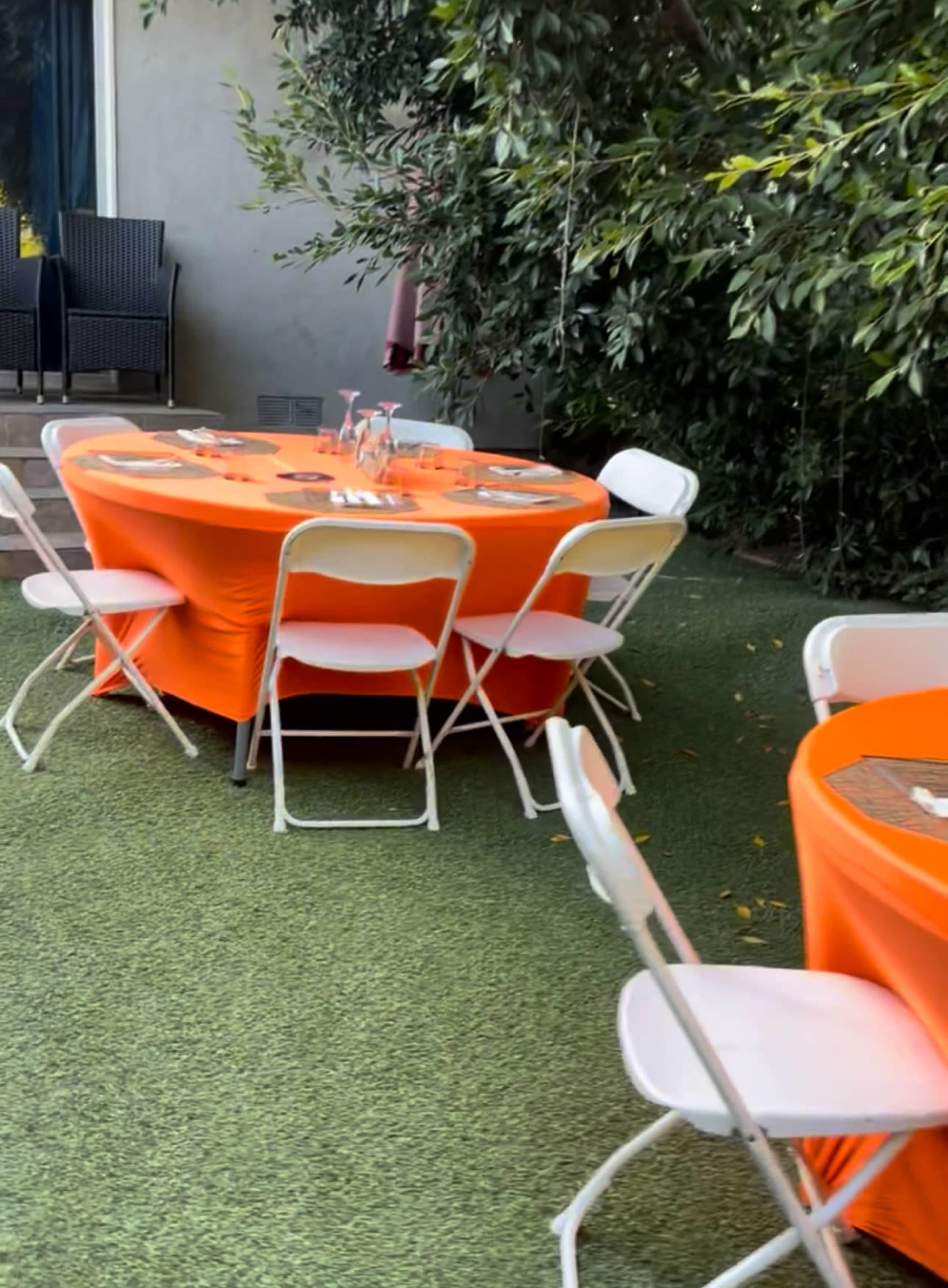 Two round tables covered in orange tablecloths are set in a green outdoor space, surrounded by white folding chairs.