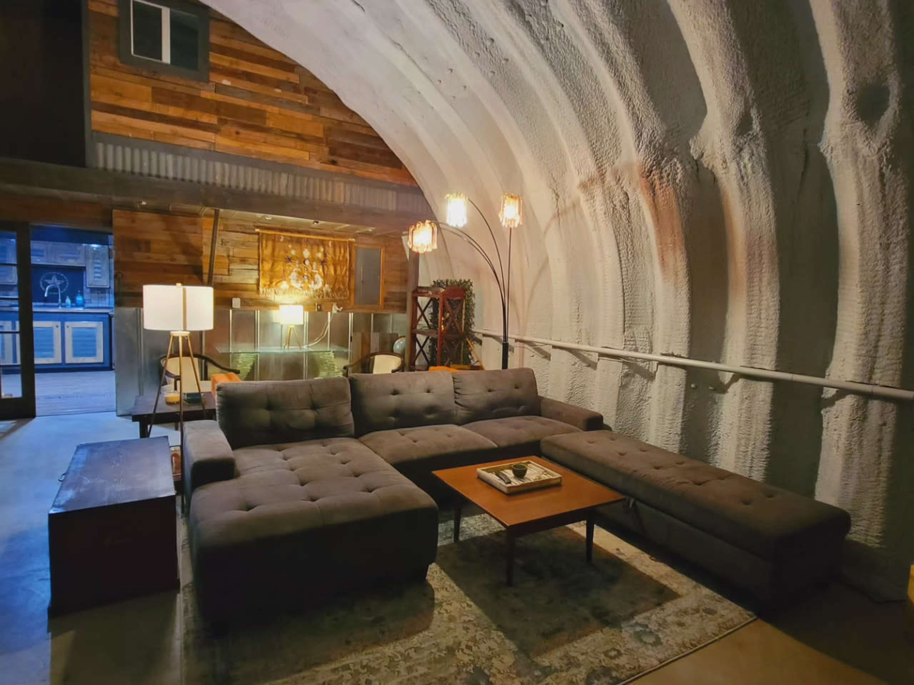 The scene features a modern living room with a large sectional sofa, a wooden coffee table, and a soft-lit ambiance created by a floor lamp and decorative light fixtures in a curved, textured space.