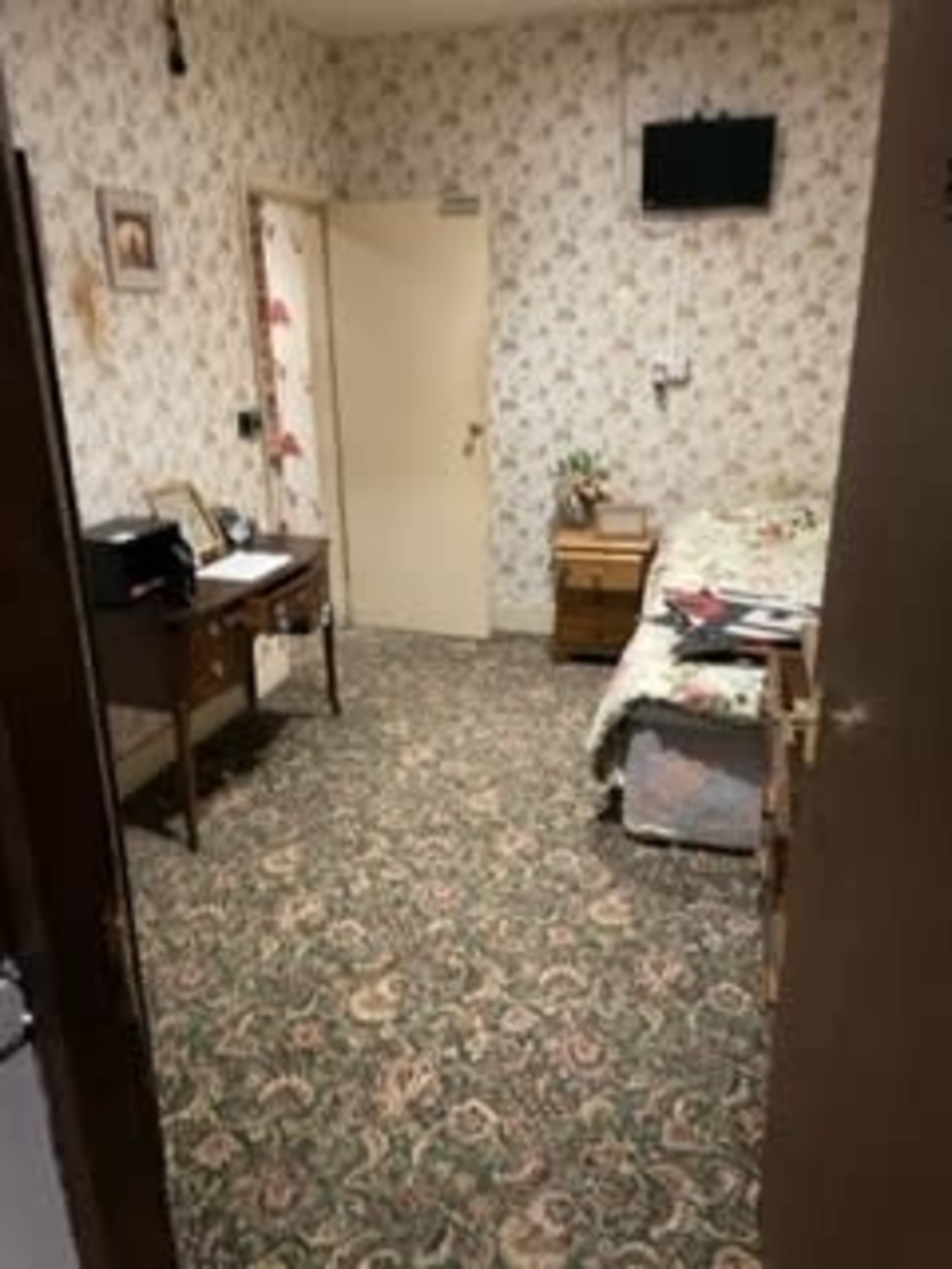 A small room with floral wallpaper, a bed covered with a patterned blanket, a desk with a chair, and a wall-mounted television.