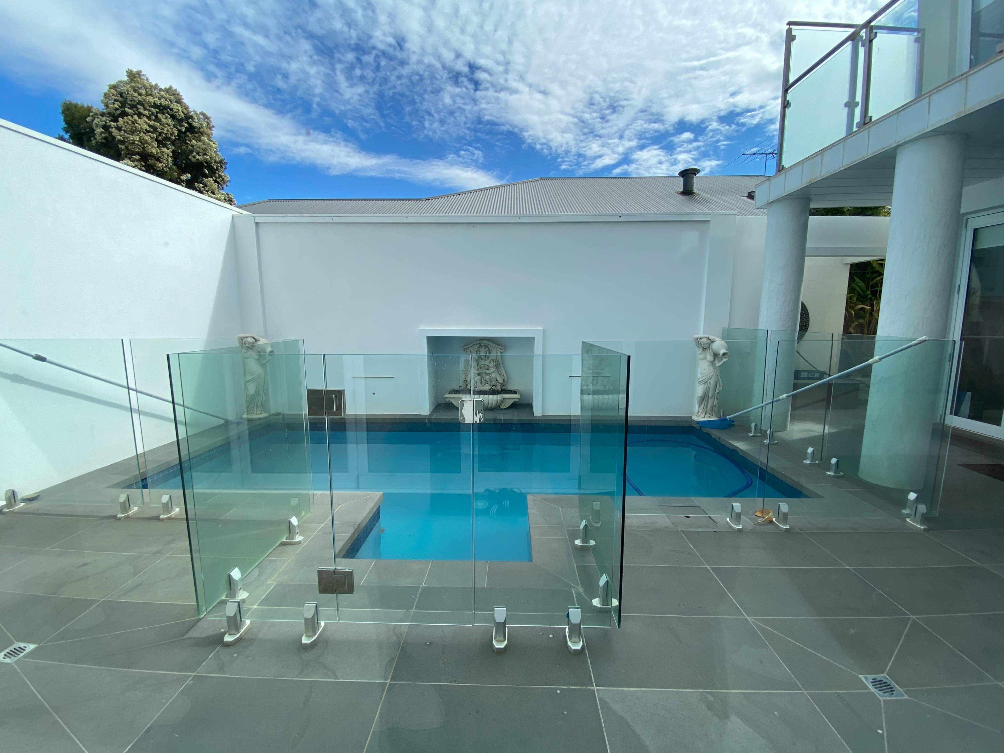 The image shows a modern pool area with glass fencing, a water feature, and a clear blue sky overhead.