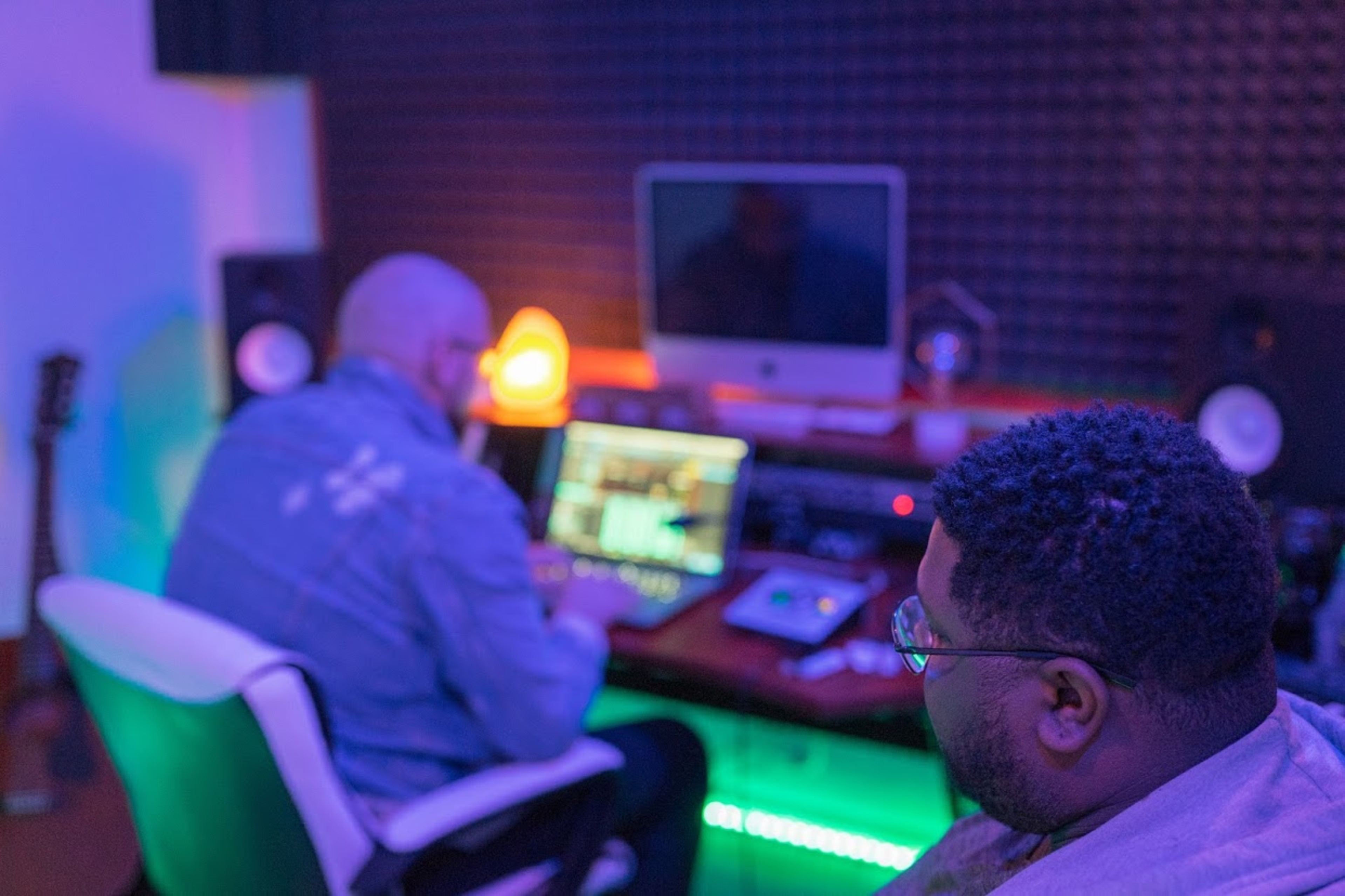 Two individuals work in a music studio, with one focused on a computer screen while the other observes, surrounded by audio equipment and ambient lighting.
