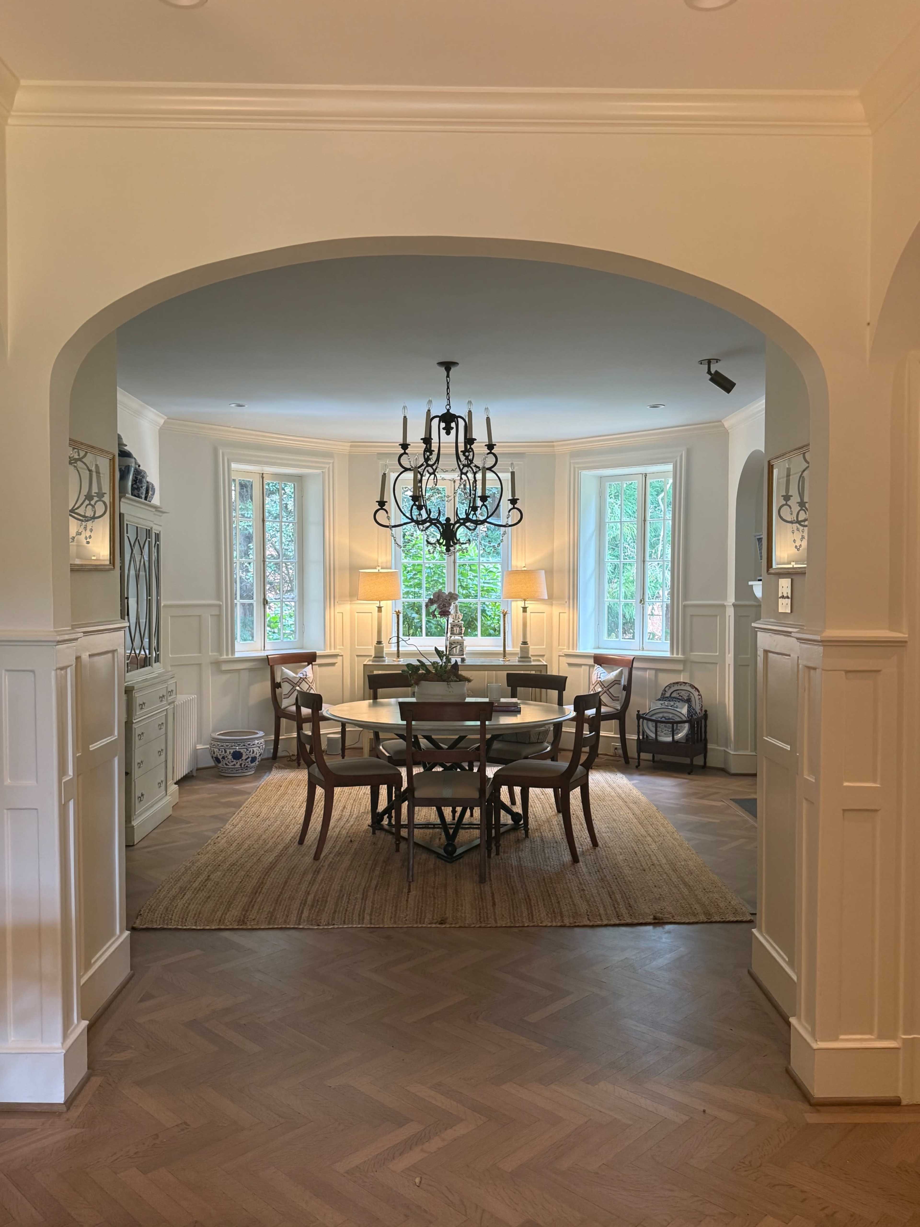 A dining area with a round table surrounded by chairs is illuminated by a chandelier, framed by archways and windows featuring greenery outside.