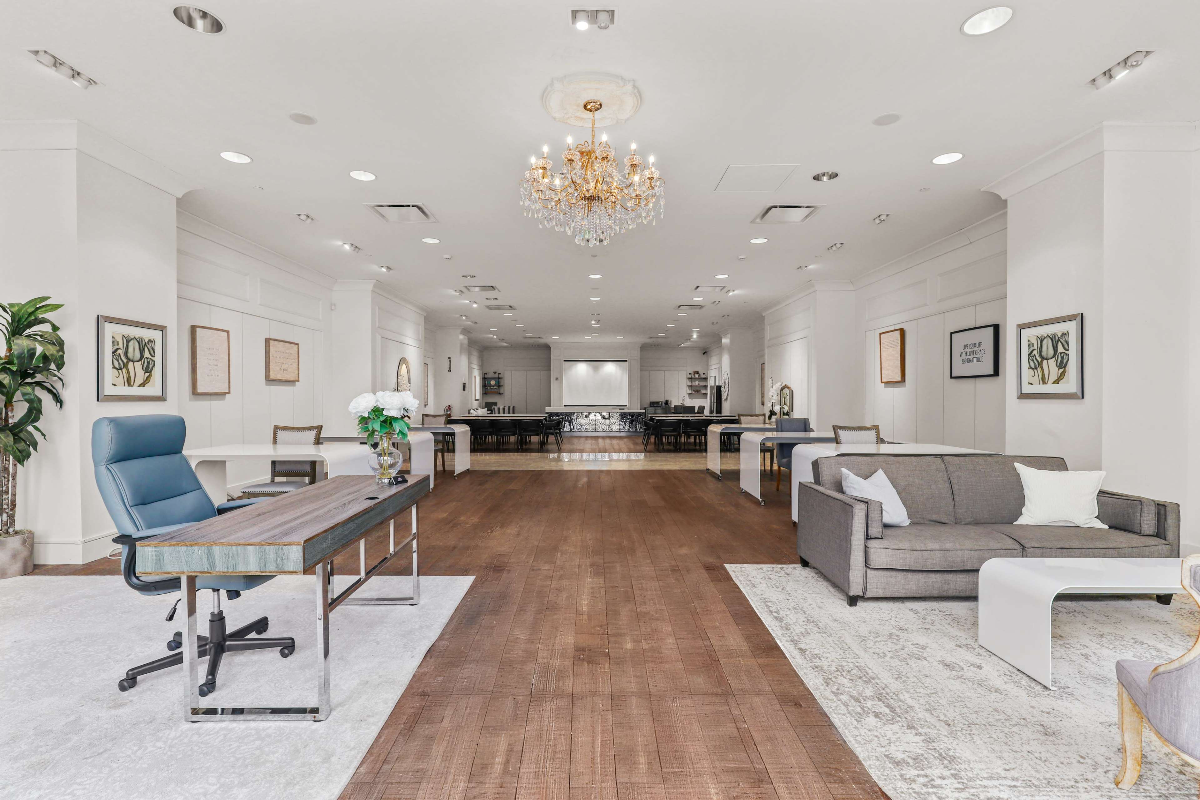 The image shows a modern, spacious lobby area featuring a desk, a sofa, a chandelier, and several tables arranged for gatherings.