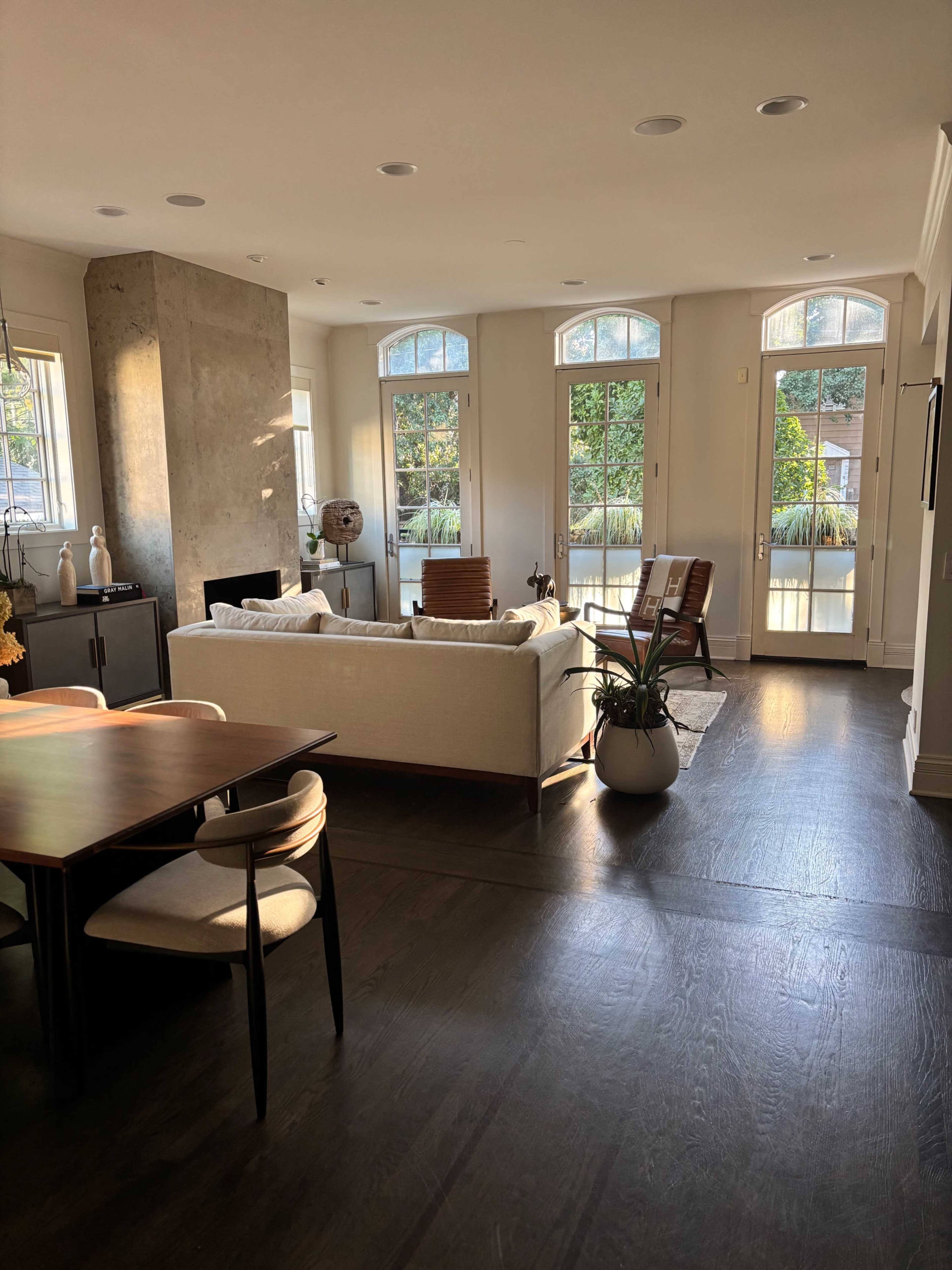 A spacious living area features large windows, a light-colored sofa, a wooden dining table, and plants, with natural light illuminating the room.