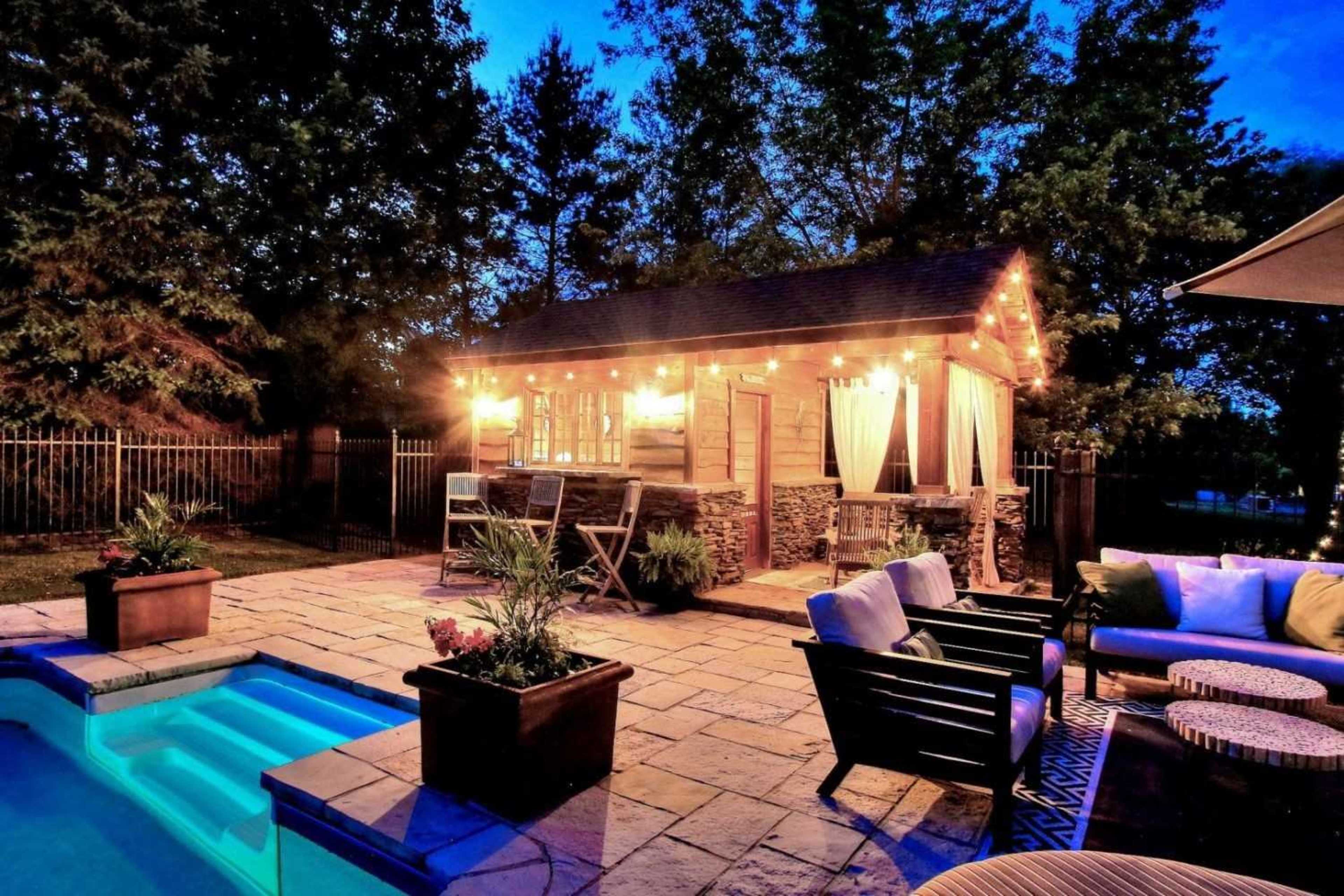 The scene features a cozy outdoor patio area with a small illuminated gazebo, a pool with blue lighting, and comfortable seating arranged around potted plants.