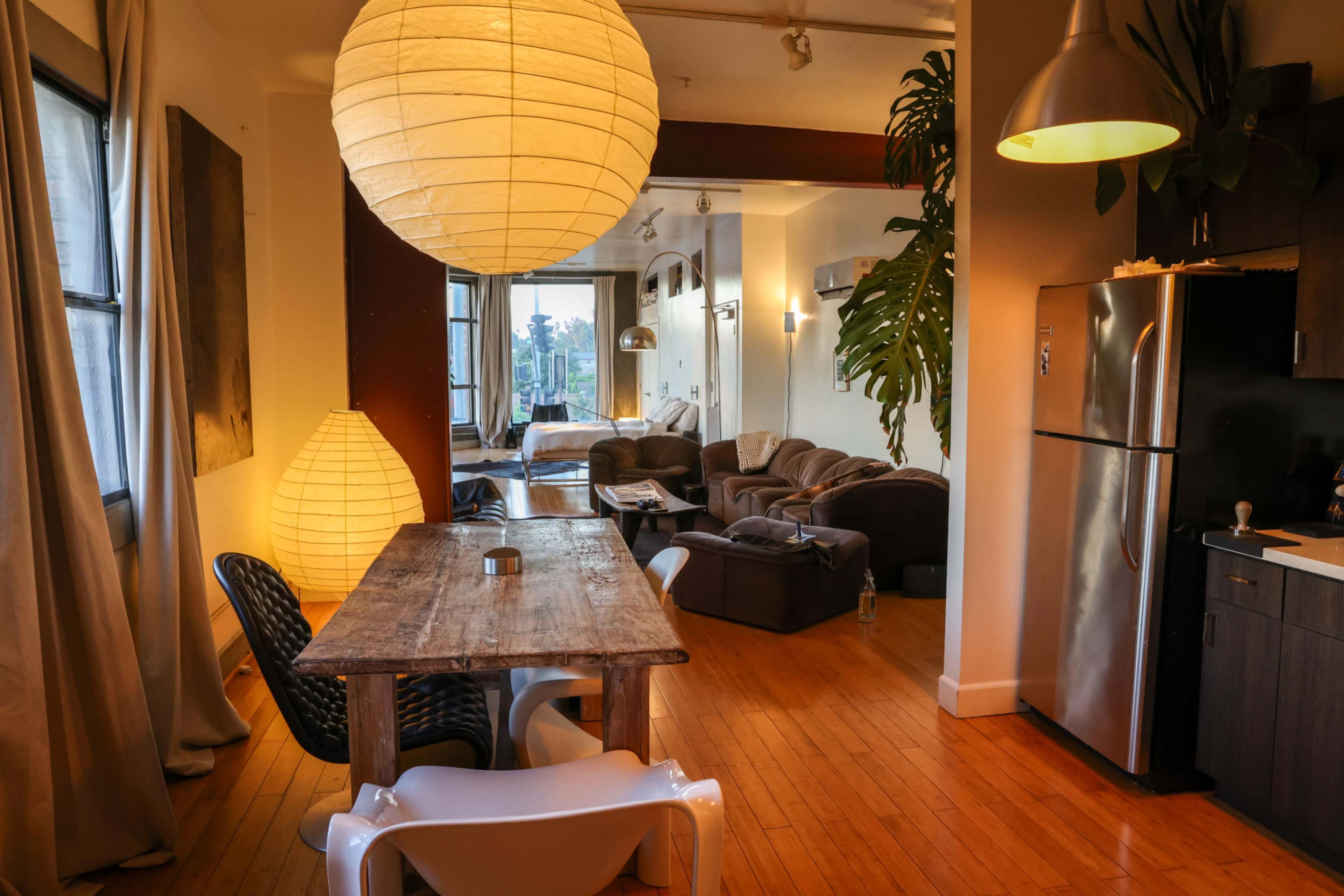 The image shows a modern interior space featuring a dining table with chairs, a large light fixture, and an open living area with plush sofas and large windows.