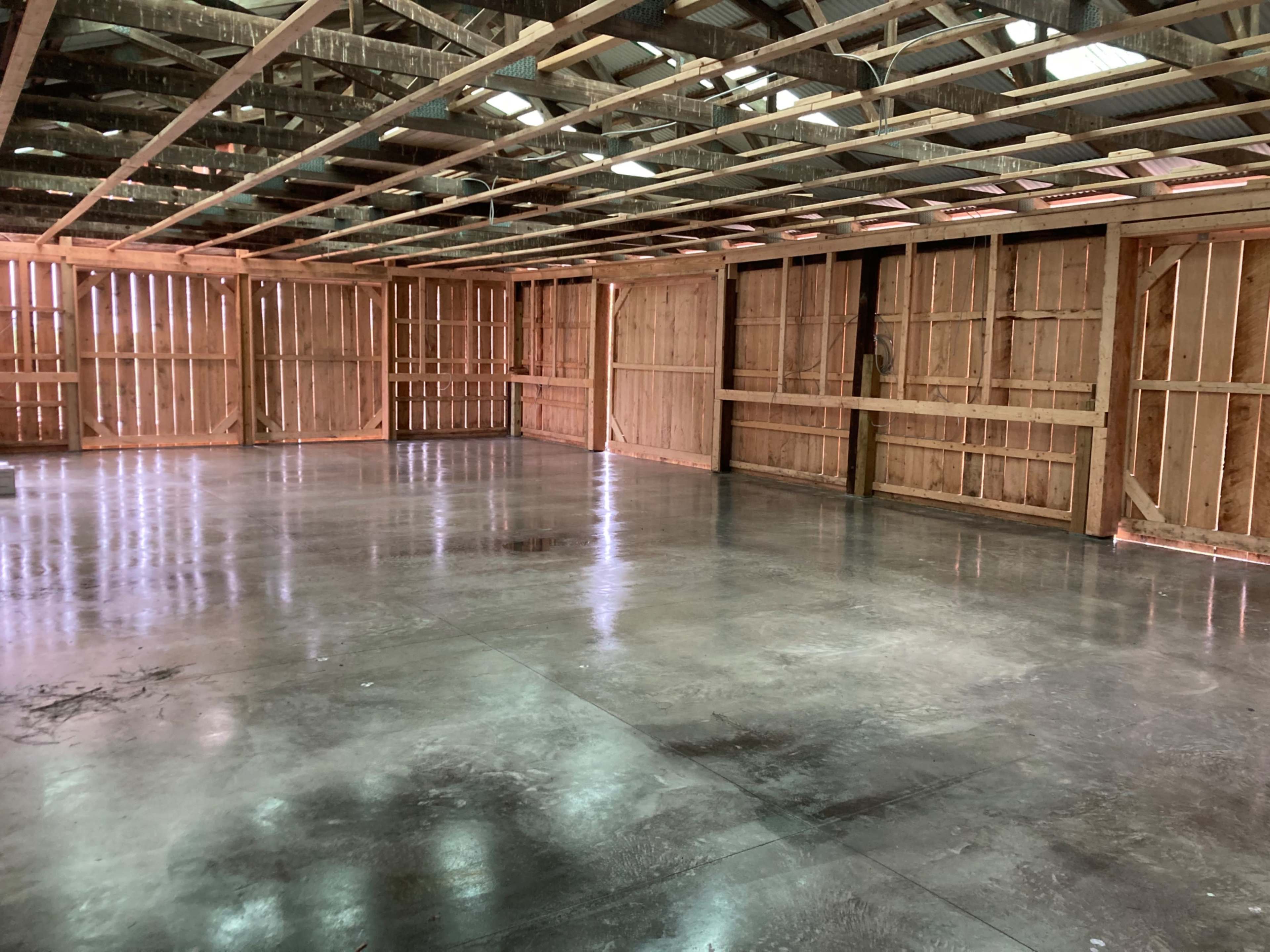 The image shows the interior of a large, unfinished wooden structure with an exposed beam ceiling and a smooth concrete floor.