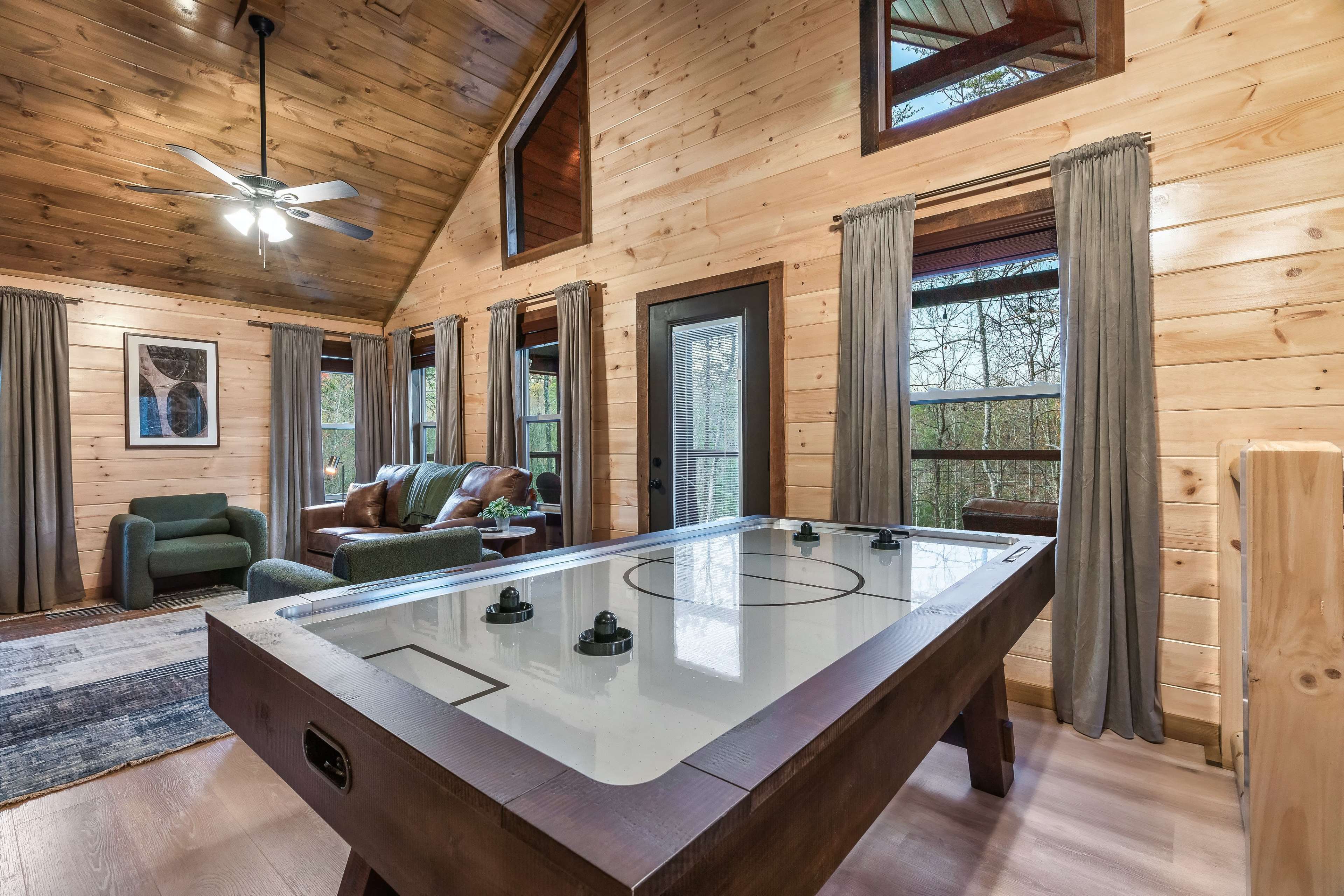 The image depicts a cozy cabin interior featuring a large air hockey table in the center, surrounded by a seating area with a couch and armchair, all set against wood-paneled walls and large windows.