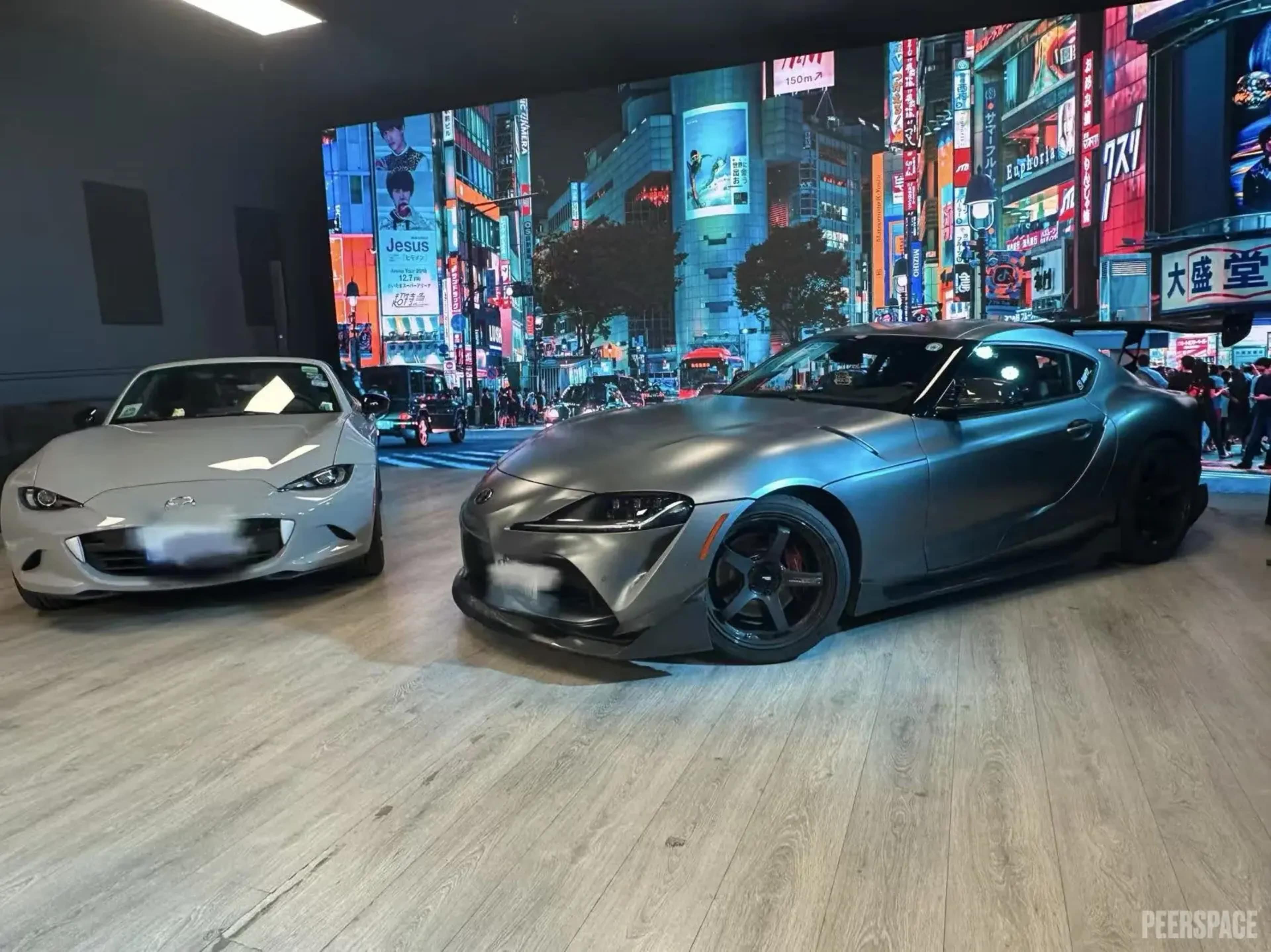 A silver sports car and a gray sports car are parked in front of a brightly lit cityscape backdrop featuring neon signs.