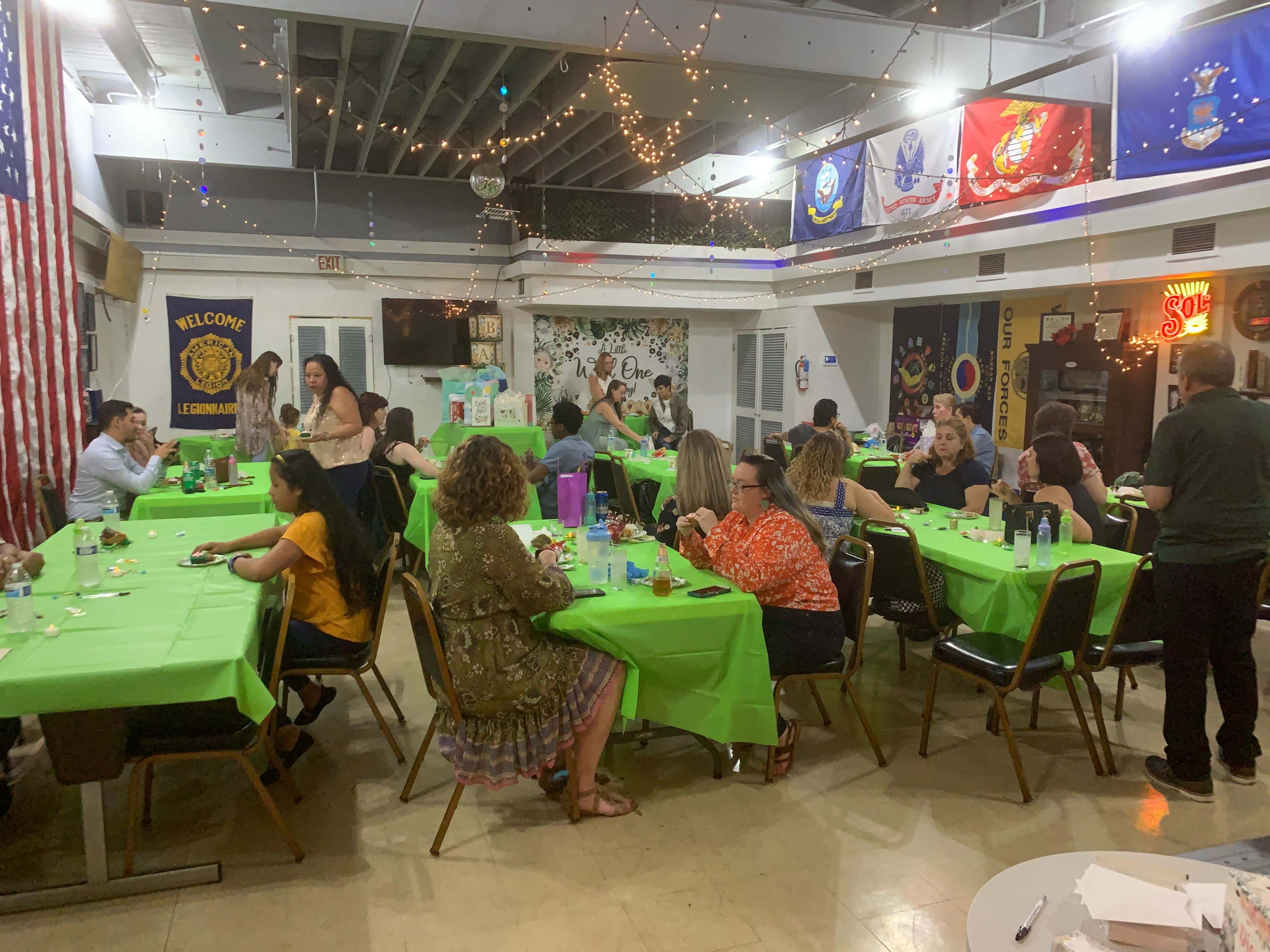 A group of people sits at tables covered with green tablecloths, enjoying a gathering in a decorated hall.