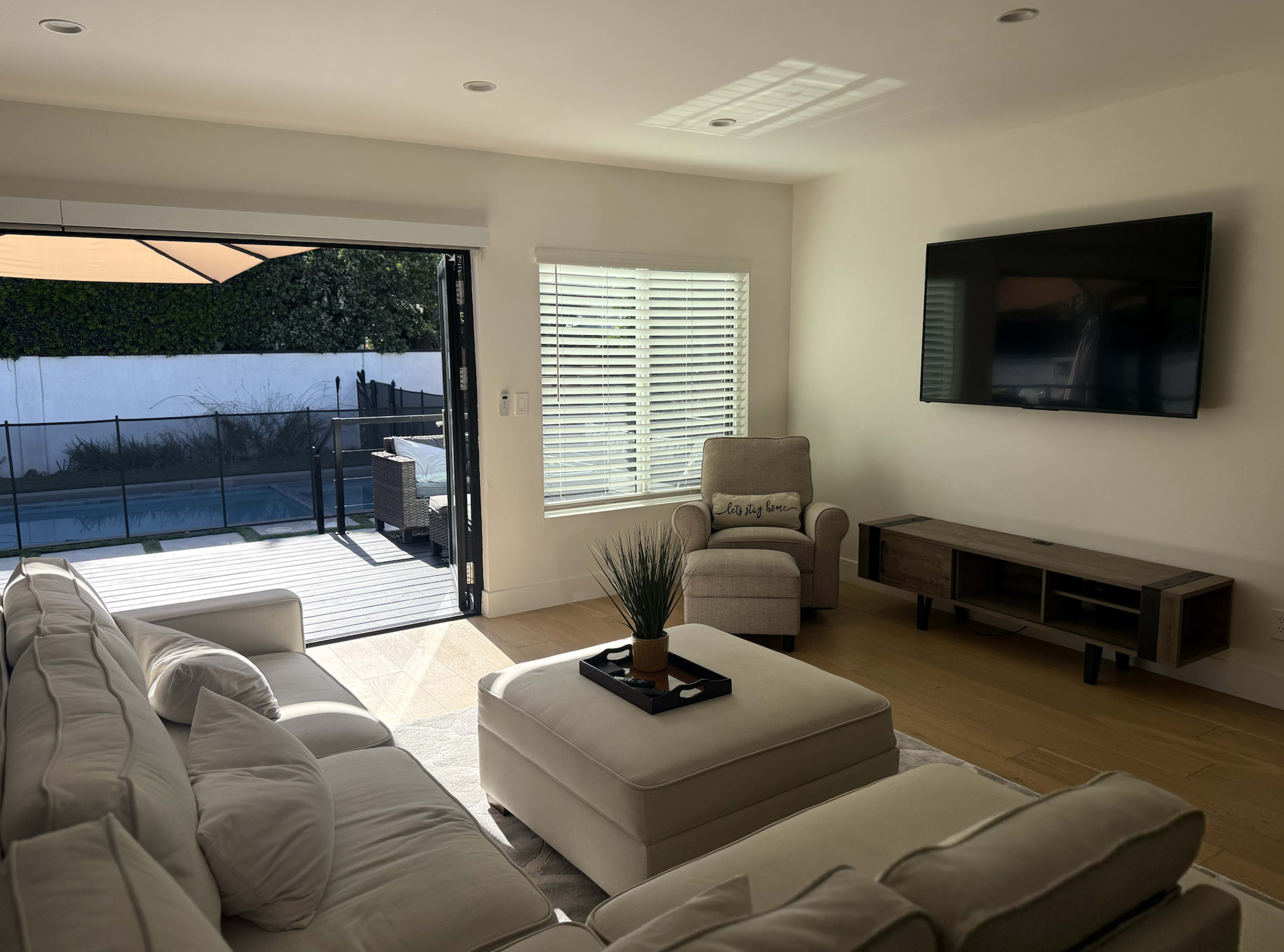 The image shows a bright living room with a large sectional sofa, an ottoman, a television mounted on the wall, and a sliding door leading to an outdoor area with a pool.
