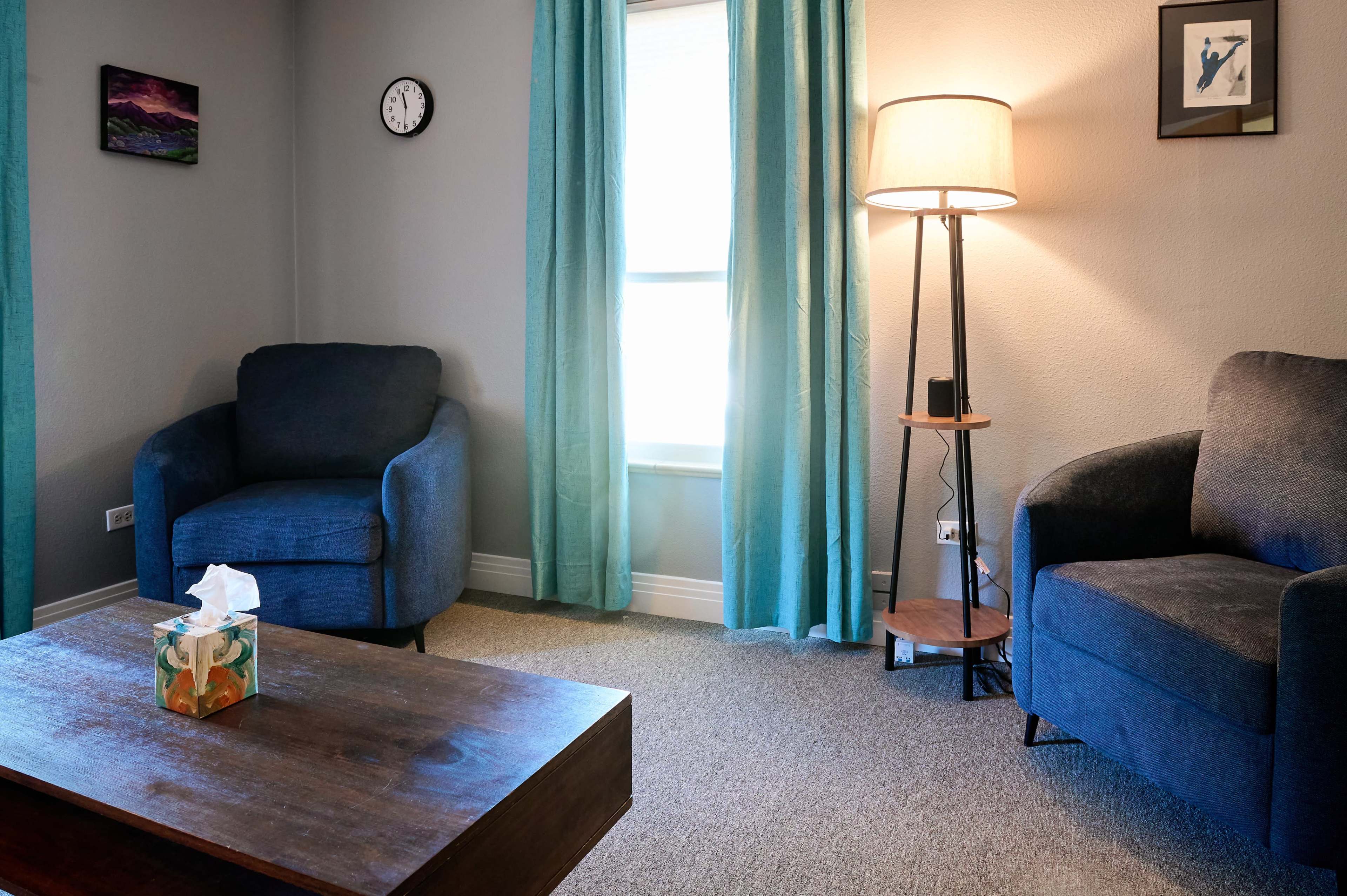 The image shows a cozy living room with two blue armchairs, a wooden coffee table with a tissue box, a standing lamp, a clock on the wall, and light blue curtains.
