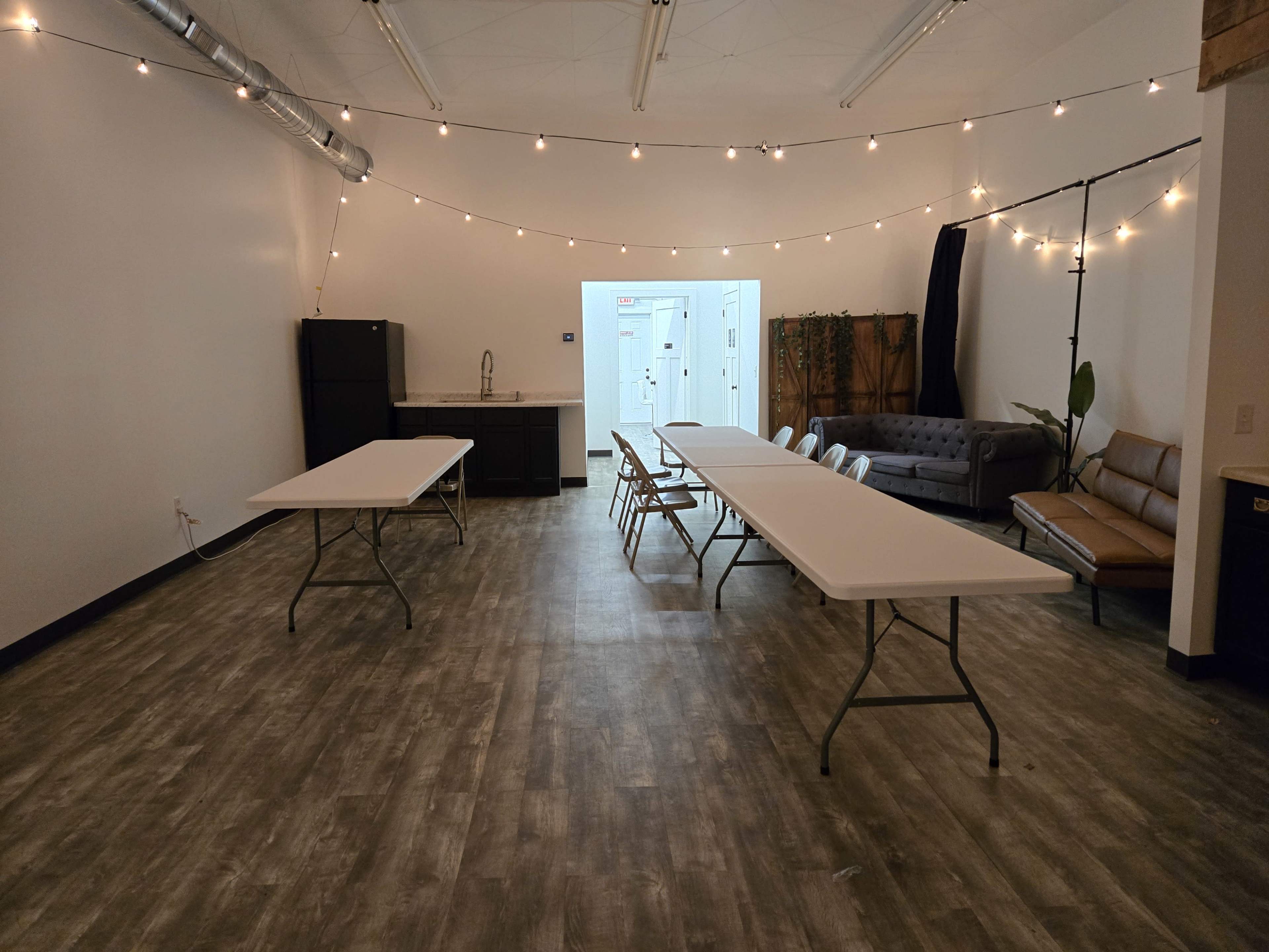 The image shows a spacious room with several folding tables and chairs arranged in the center, string lights overhead, and a kitchenette area on the side.