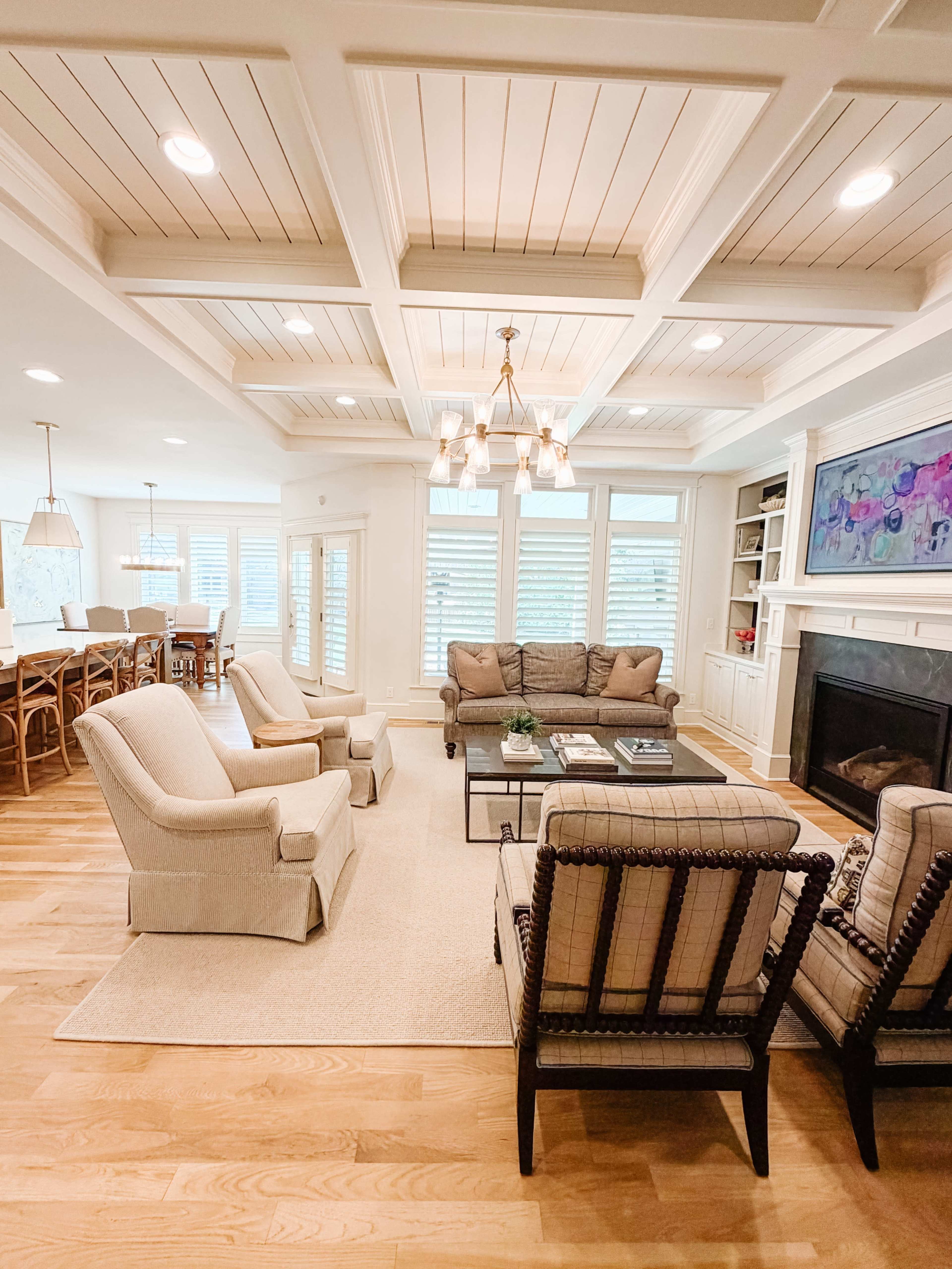 The image shows a spacious living room with a coffered ceiling, featuring beige sofas and chairs arranged around a glass coffee table, alongside a fireplace and large windows allowing natural light.