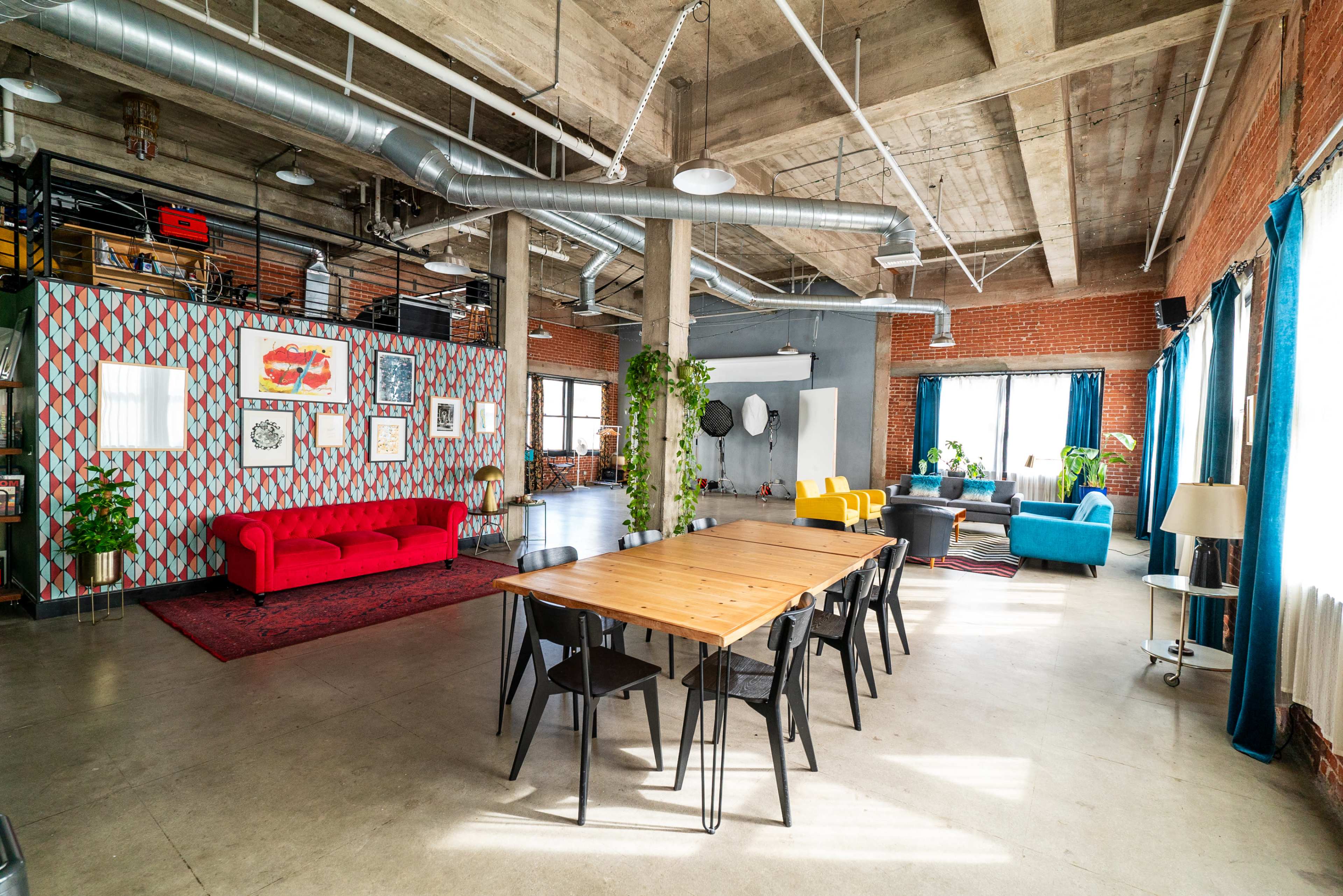 The image shows a spacious industrial-style loft featuring a colorful seating area, a long wooden dining table, and large windows providing natural light.