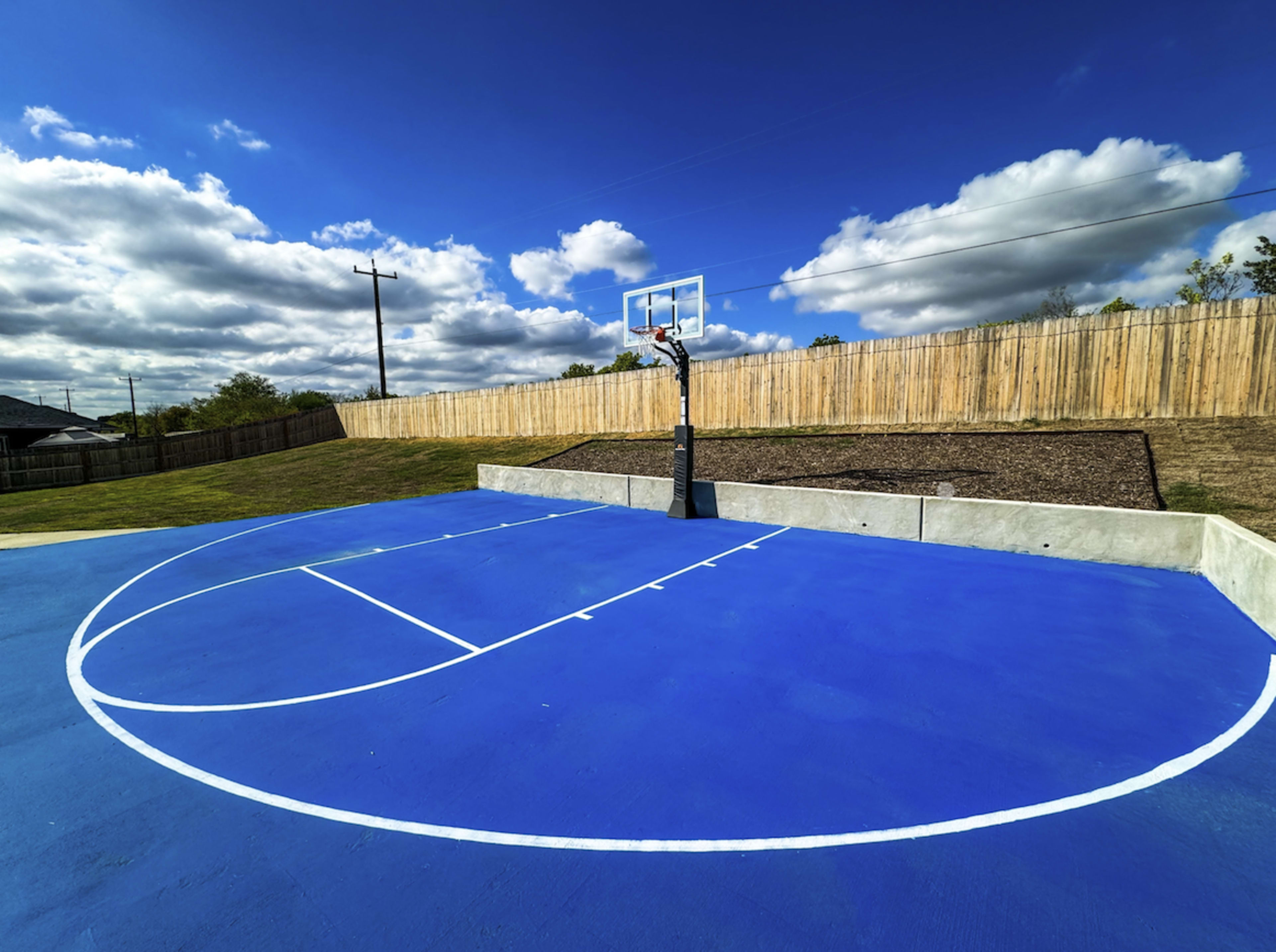 The image shows a blue basketball court with a hoop, set against a backdrop of cloudy skies and a wooden fence.