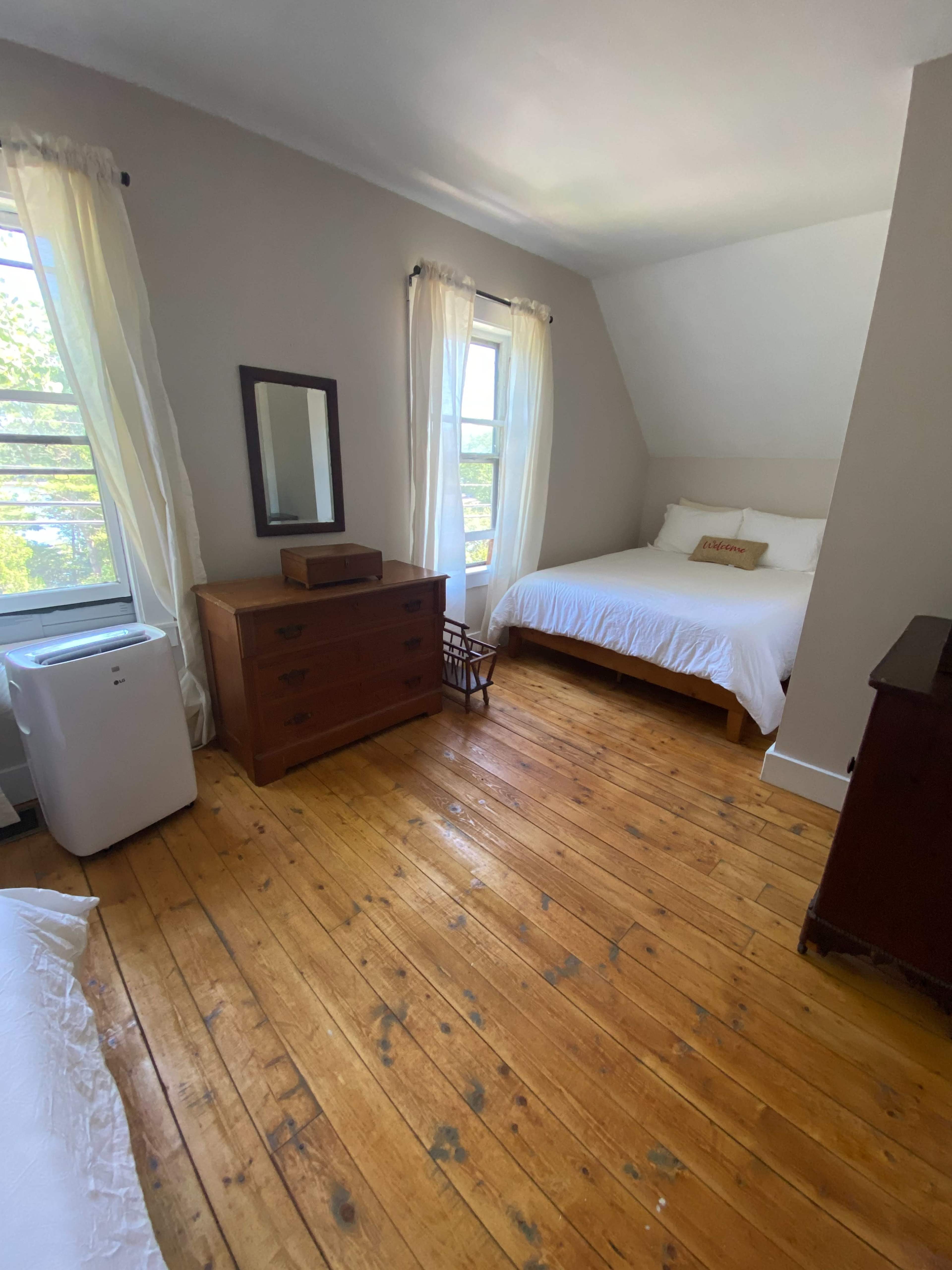 A bedroom with a wooden floor, a bed with white bedding, a wooden dresser, and two windows with sheer curtains.