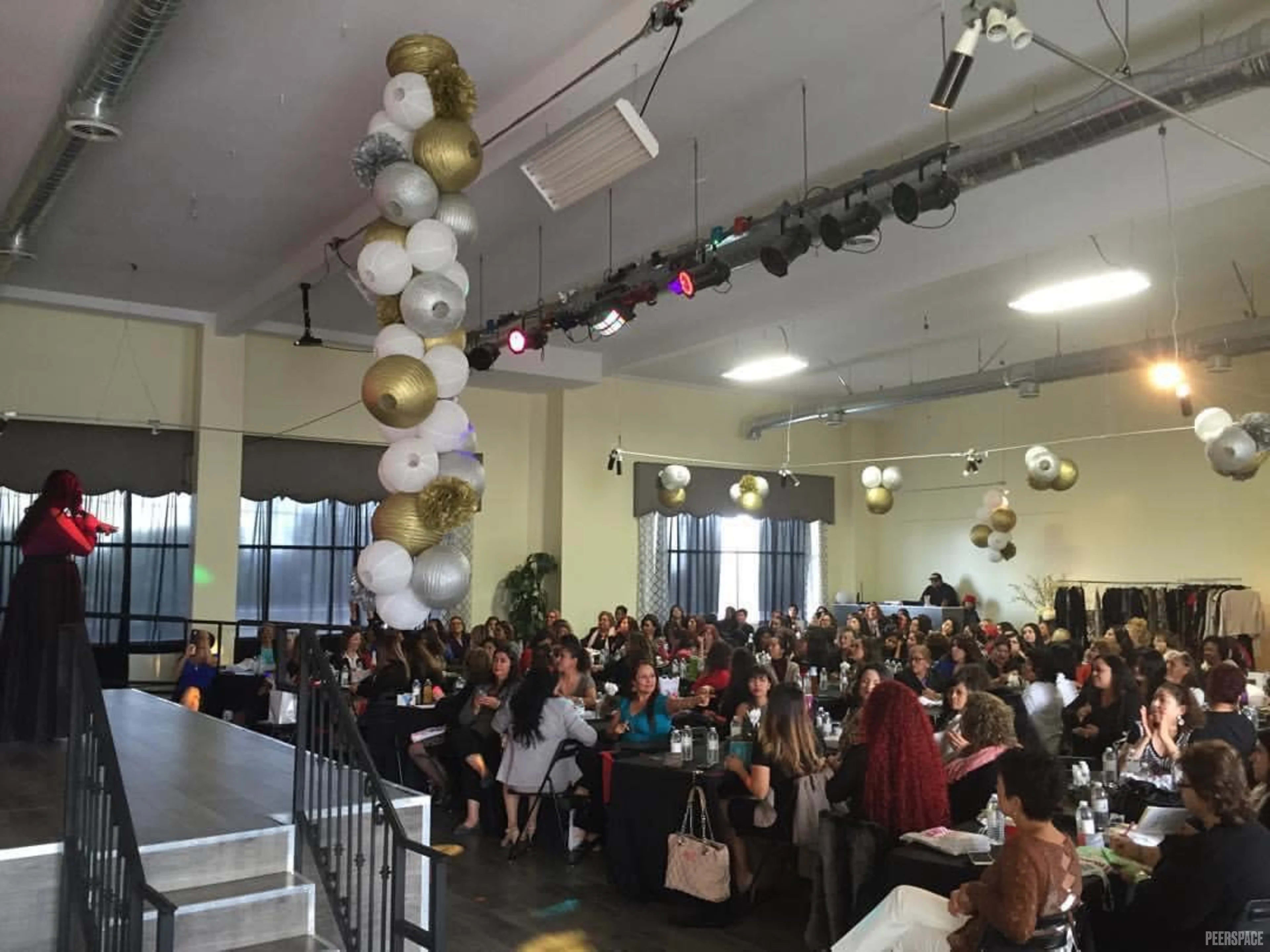 A speaker stands on a stage in front of a large audience seated at tables in a well-lit room adorned with decorative balloons.