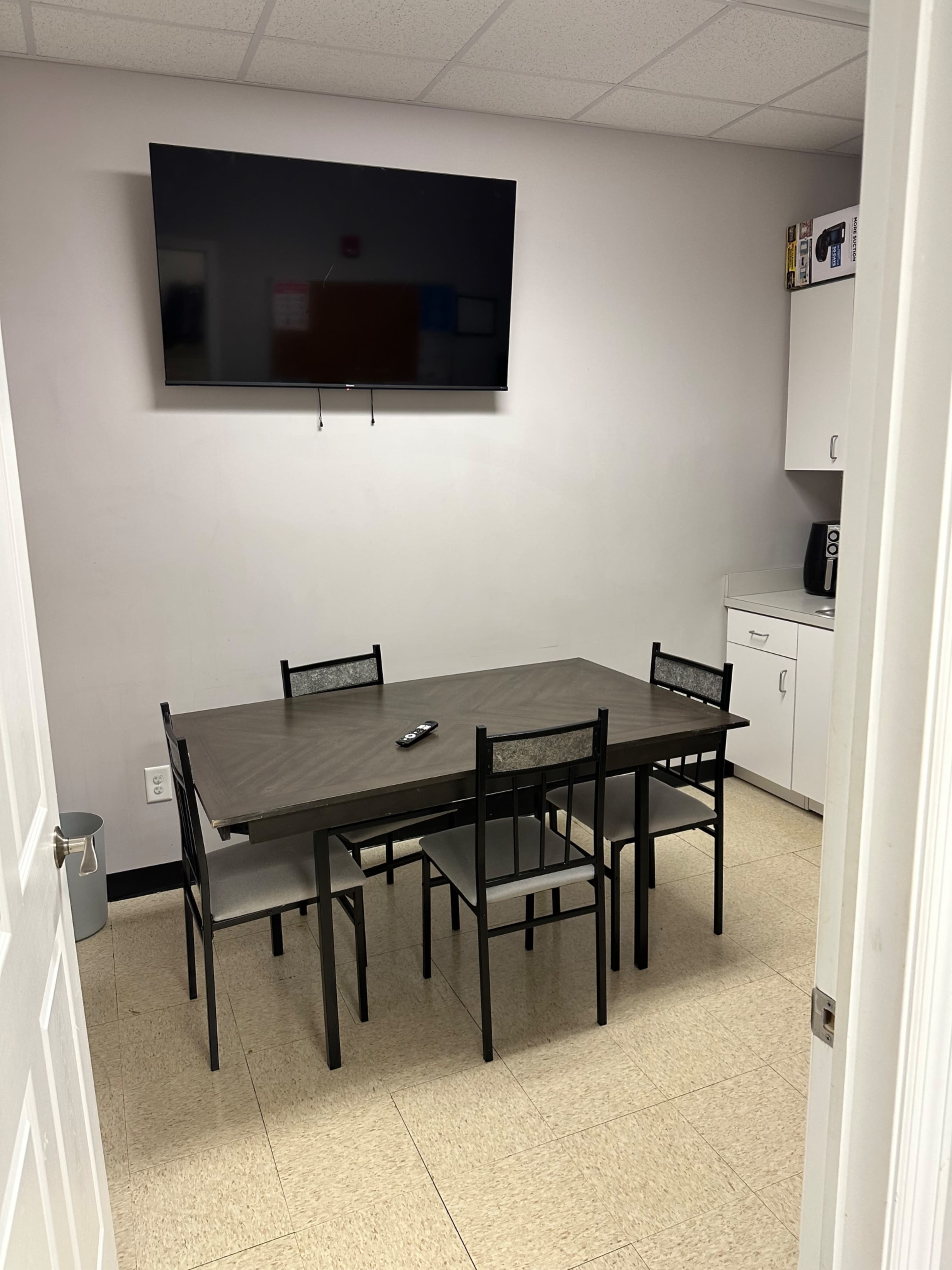 A small break room with a table and four chairs, a wall-mounted TV, and a kitchenette area.