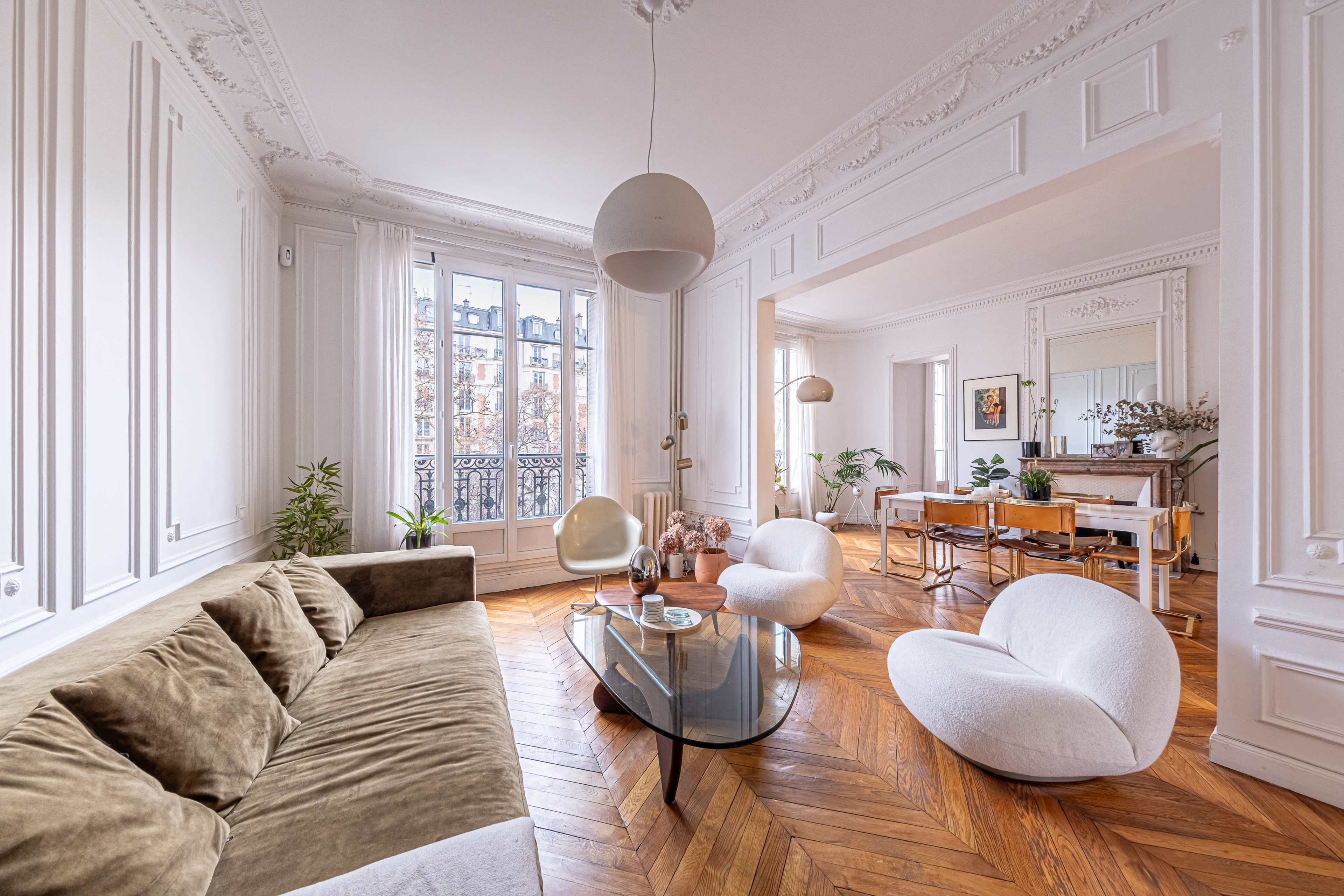 The image shows a stylish interior space featuring a brown velvet sofa, minimalist white chairs, a glass coffee table, and large windows with decorative molding.