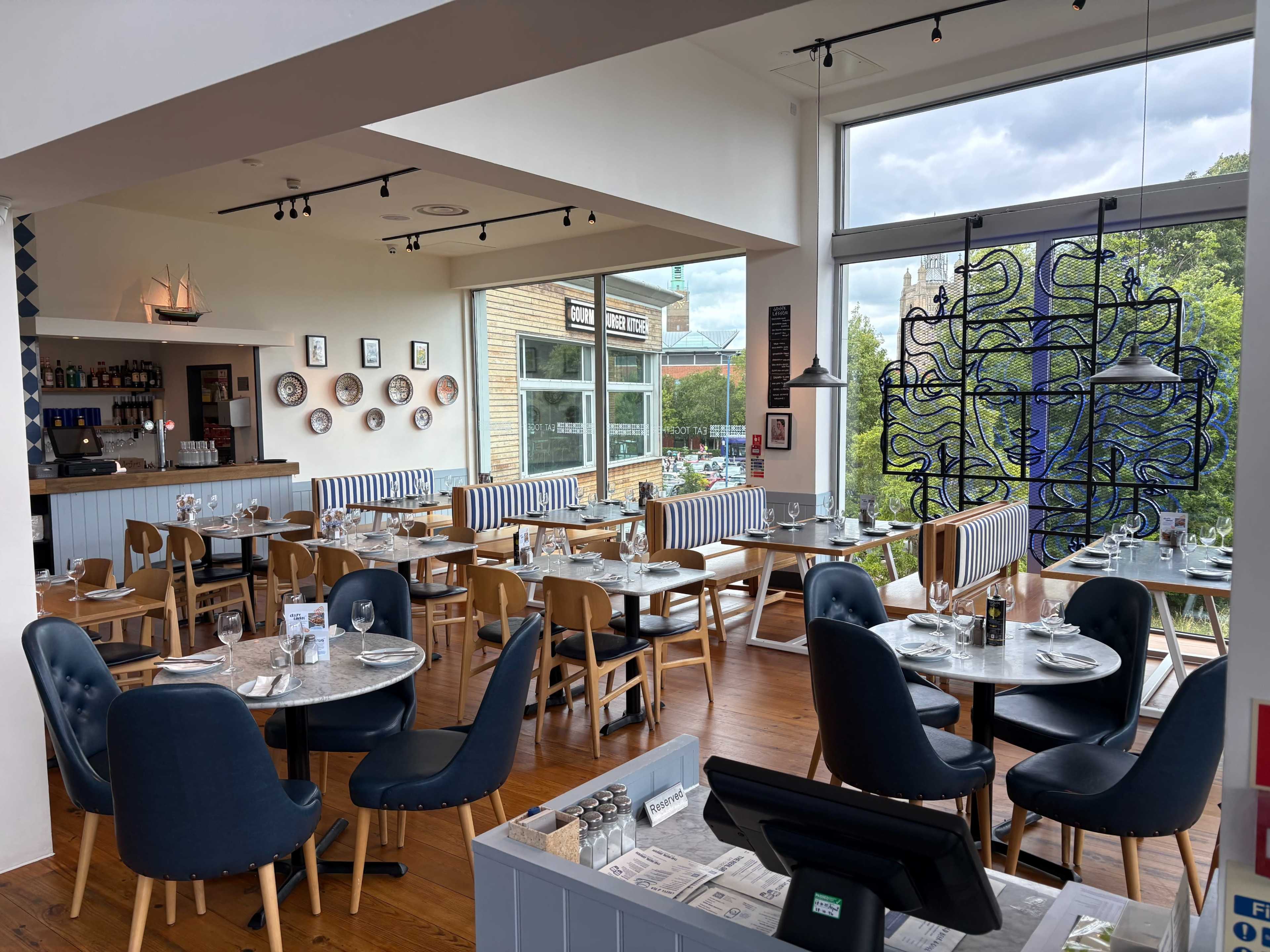 The image shows a modern restaurant interior with wooden floors, tables set for dining, and large windows overlooking an outdoor view.